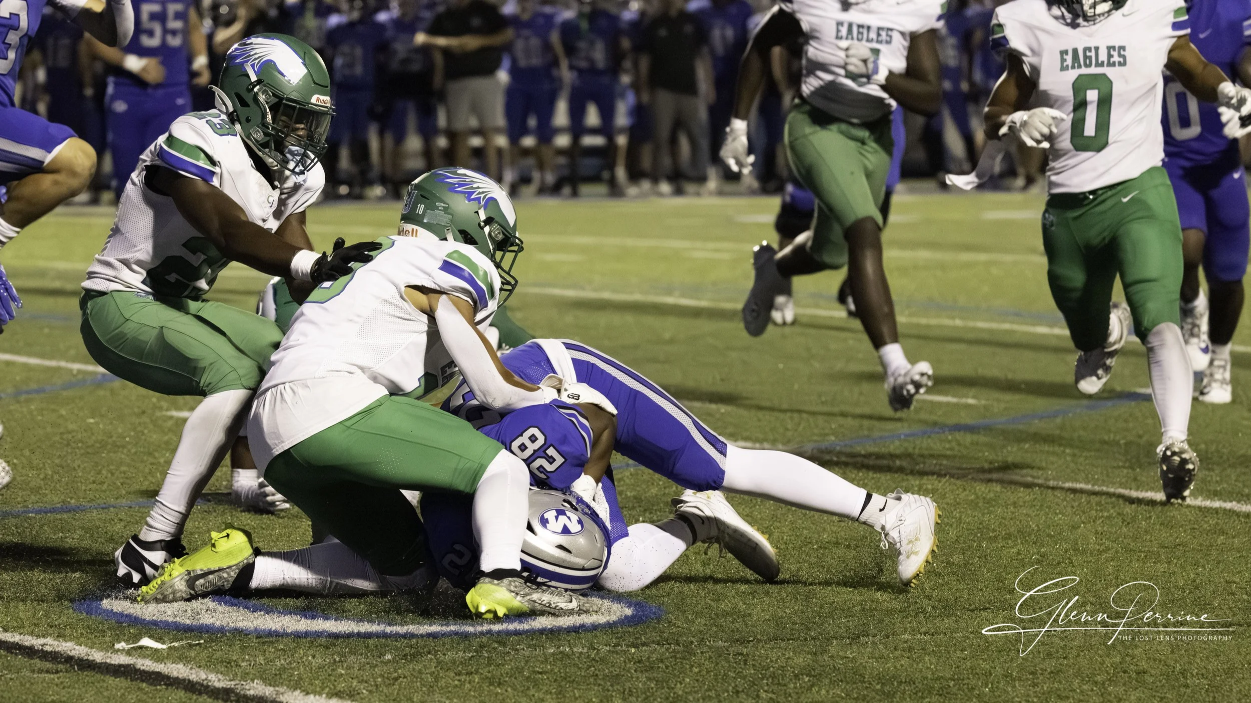 Football players from the Eagles and an opposing team are engaged in a tackle during a game on a green field at night. An Eagle player is on the ground holding the ball while other players surround him.