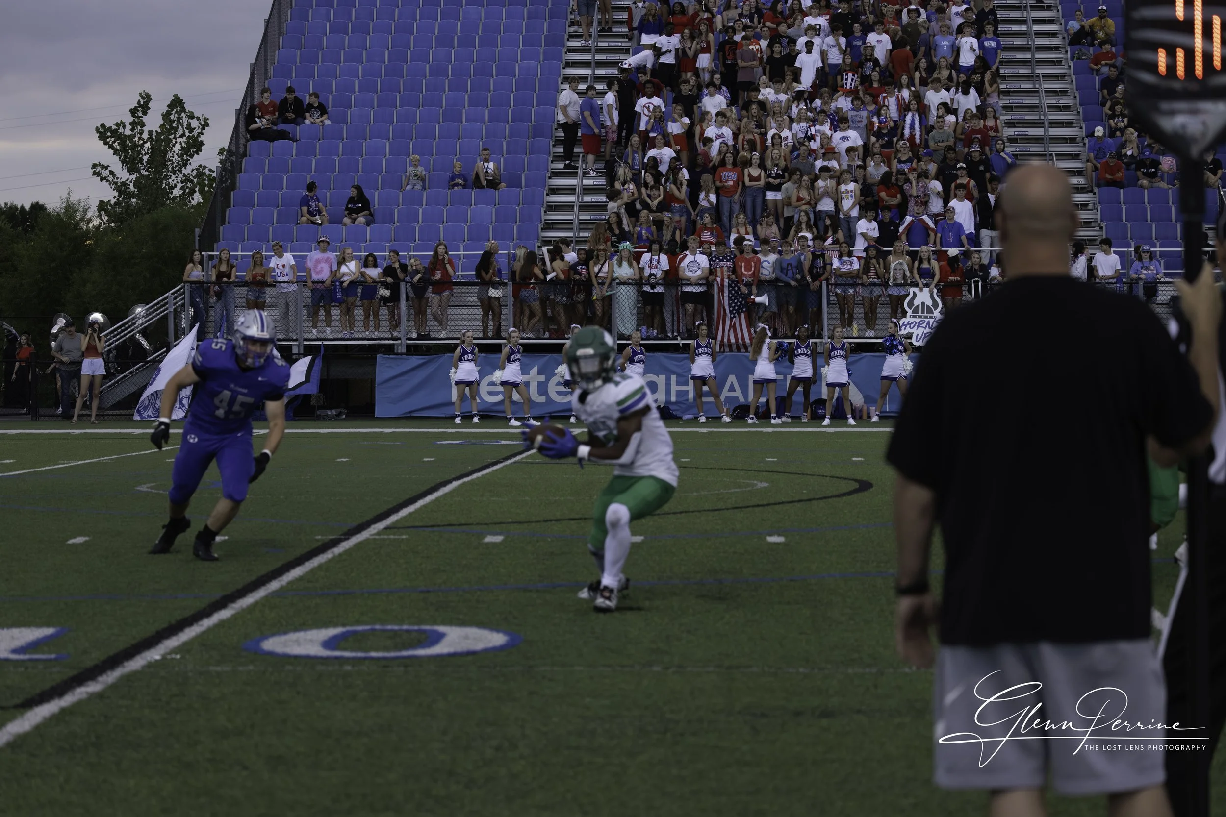 A football game in progress with two players running on the field, one in a blue uniform and the other in a white and green uniform, while a coach or staff member watches nearby, and cheerleaders are lined up on the sidelines with a crowd in the stan