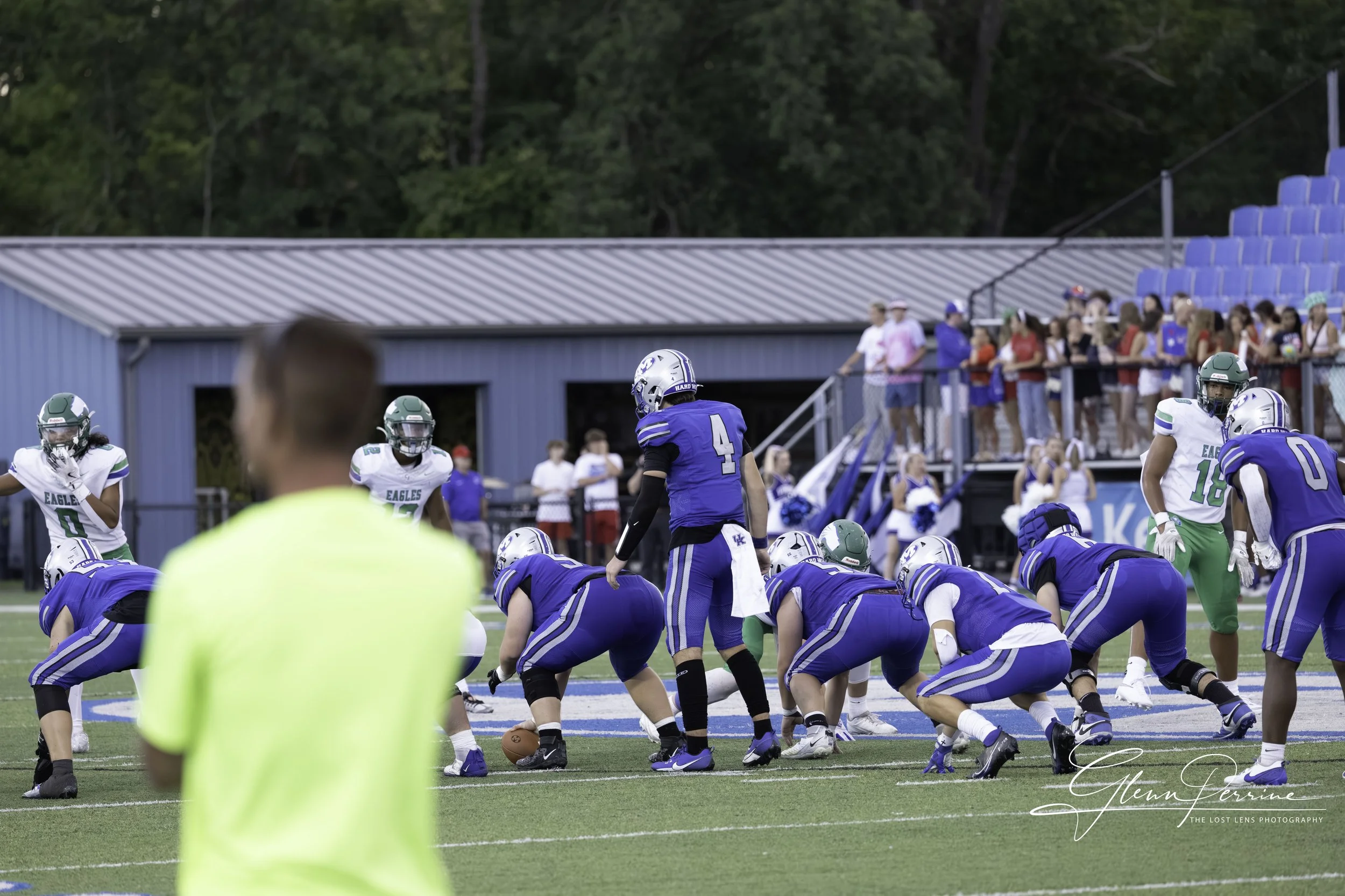 Youth American football game with players in blue and white uniforms on the field, spectators in the stands, and a coach or referee in a yellow shirt in the foreground.