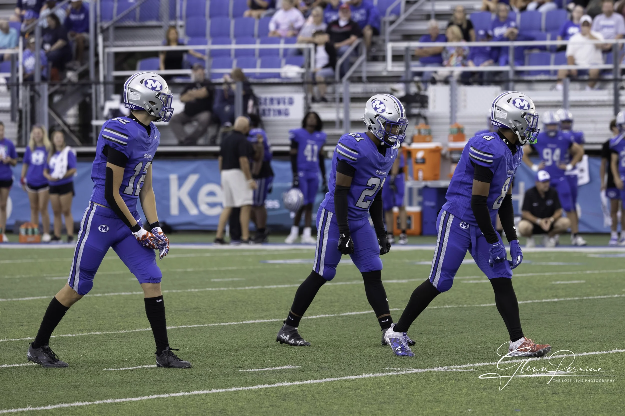 Three football players in blue jerseys and helmets are preparing for the game on a football field, with spectators and cheerleaders in the background.