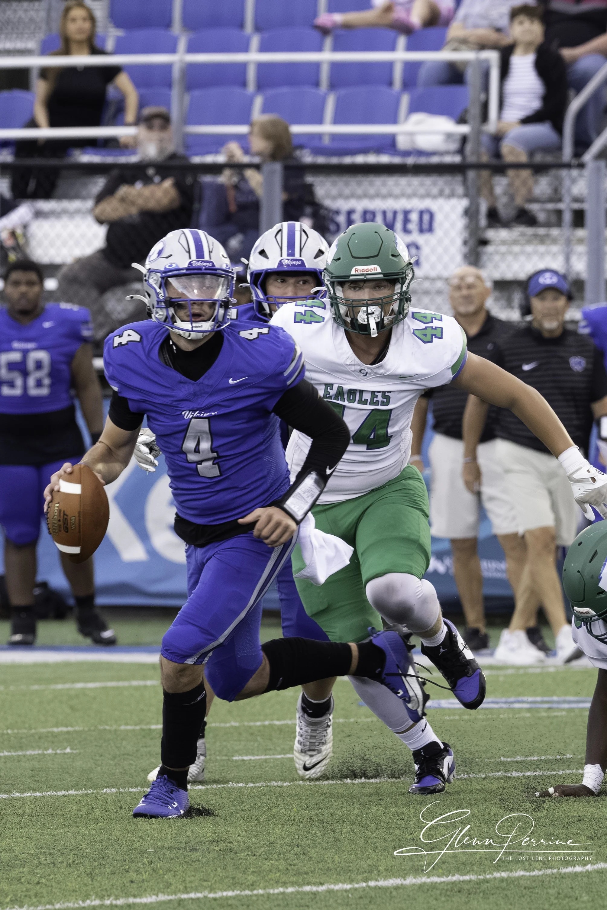 A young football player in a blue uniform with the number 4 is running with the football, evading a defender in a white and green uniform, during a game on a green field with spectators in the stands.