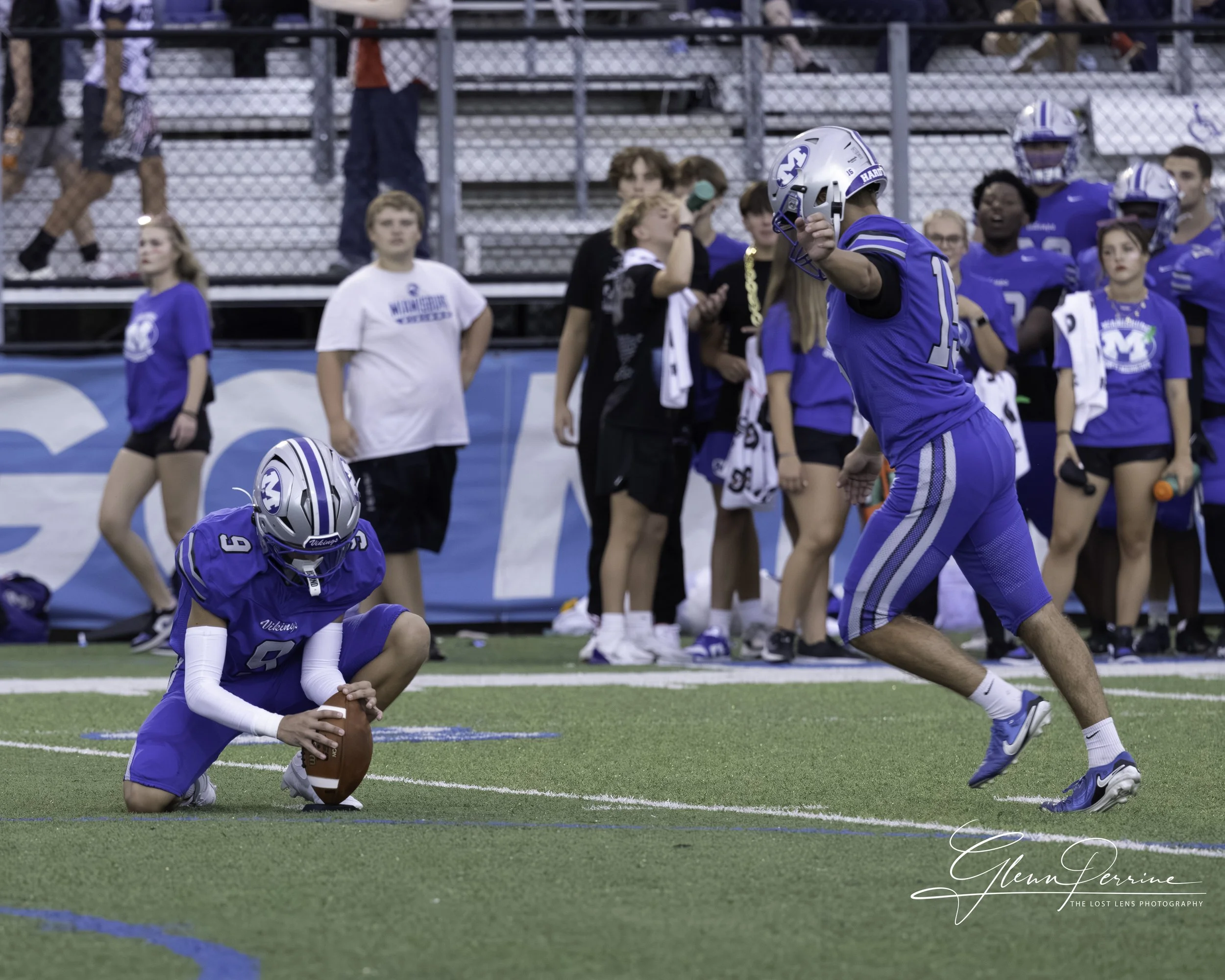 A football player in a blue uniform kneels on one knee, holding a football on the ground, while another player in a blue uniform runs towards him on the field. Spectators and team members stand in the background.