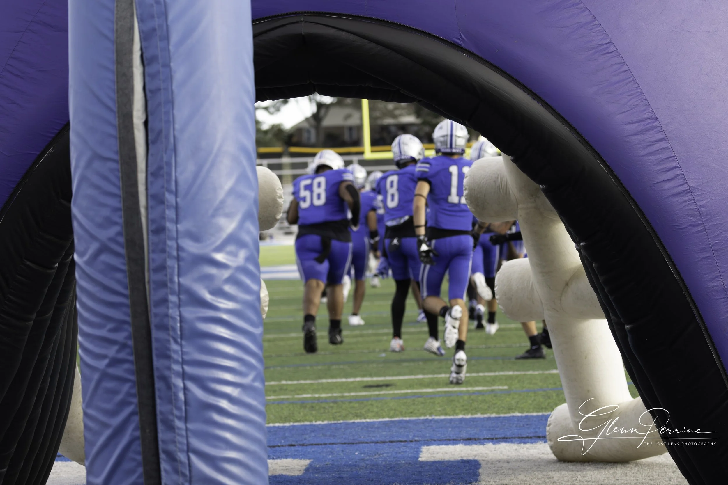 View of football players in blue uniforms running onto the field through a tunnel on a football field, with yellow goalposts visible in the background.