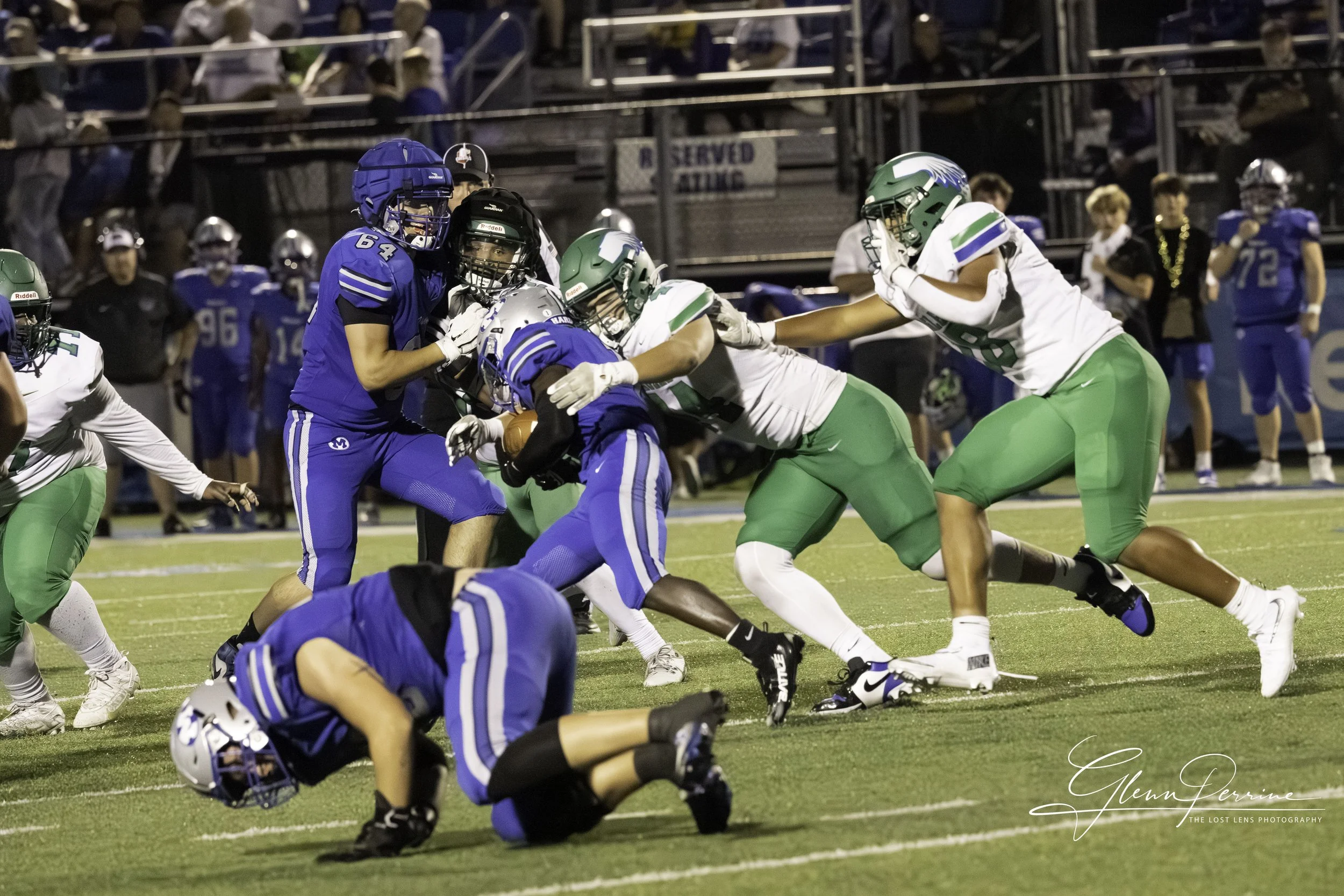Football players in blue and green uniforms engaging during a game on a field at night.