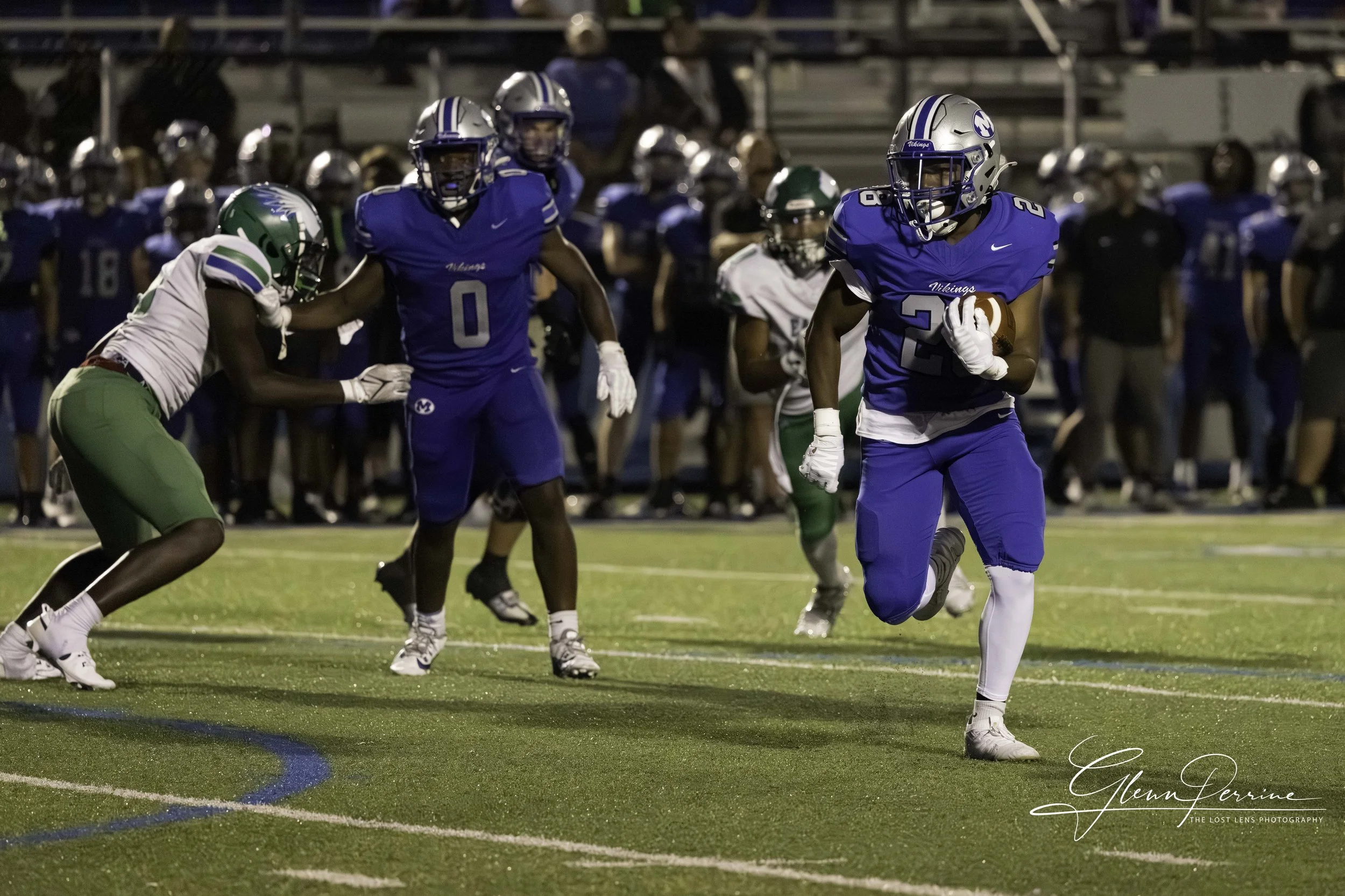 A football player in a blue uniform running with the ball on a football field during a game at night.