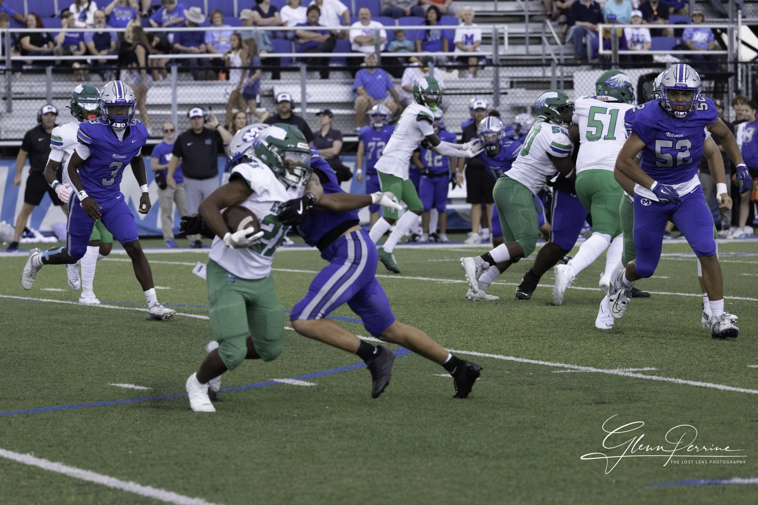 Football game with players running on field, some players in white and green uniforms and others in blue uniforms, with spectators and coaches in the background.
