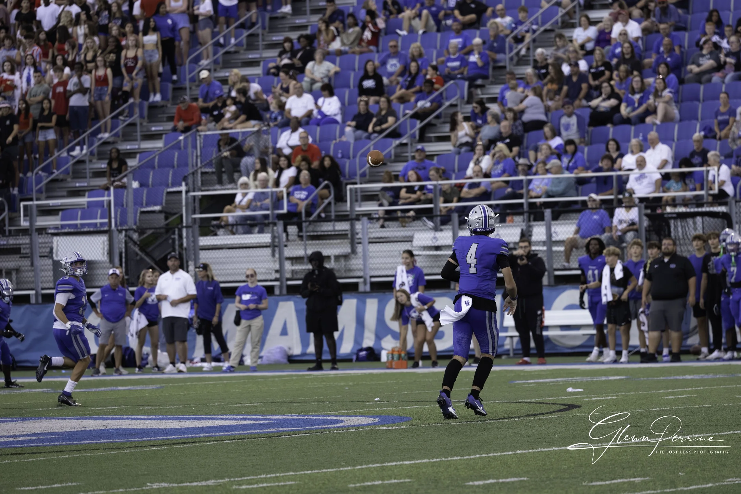 A football game with players in blue uniforms on the field, with one player about to catch a football. Spectators are seated in the stands behind the players.