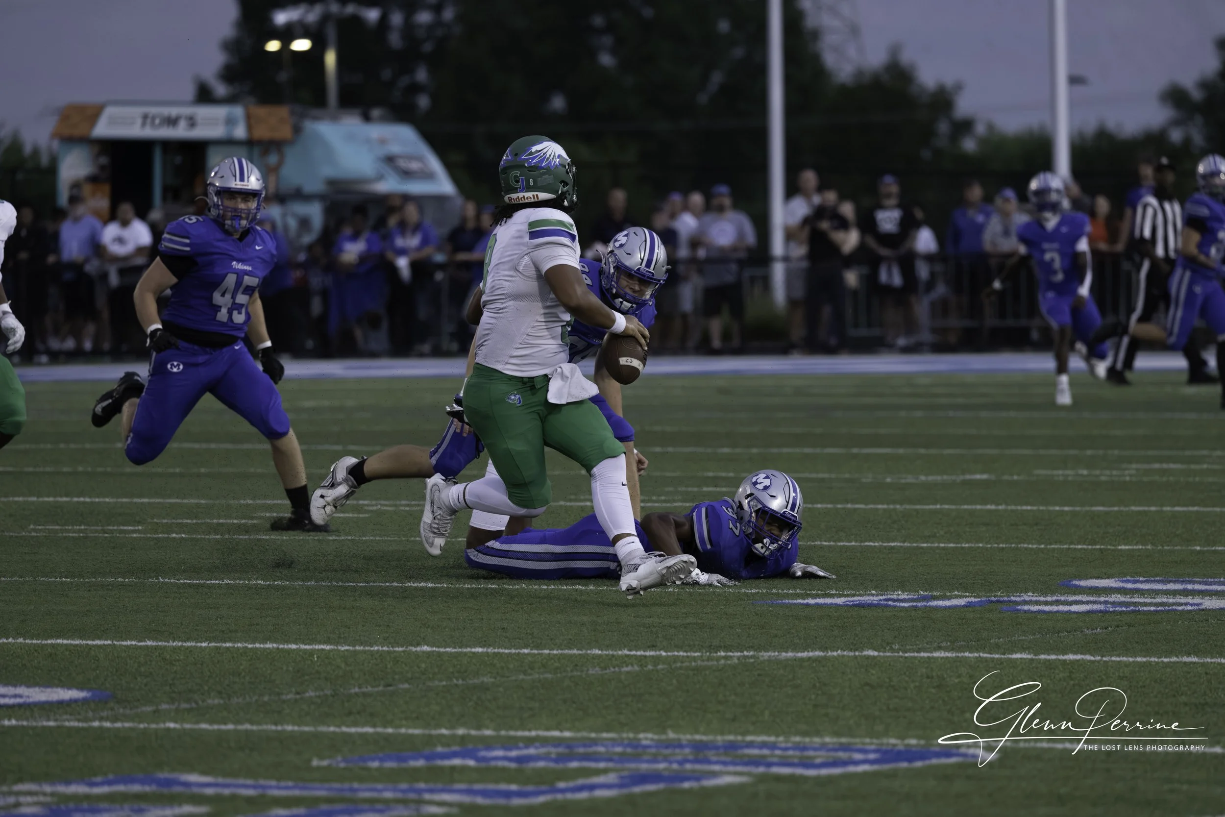 An American football game in progress with players from two teams, one in blue jerseys and the other in white and green, on the field during evening hours. A player in a green jersey is holding the football while being tackled by a player in blue who
