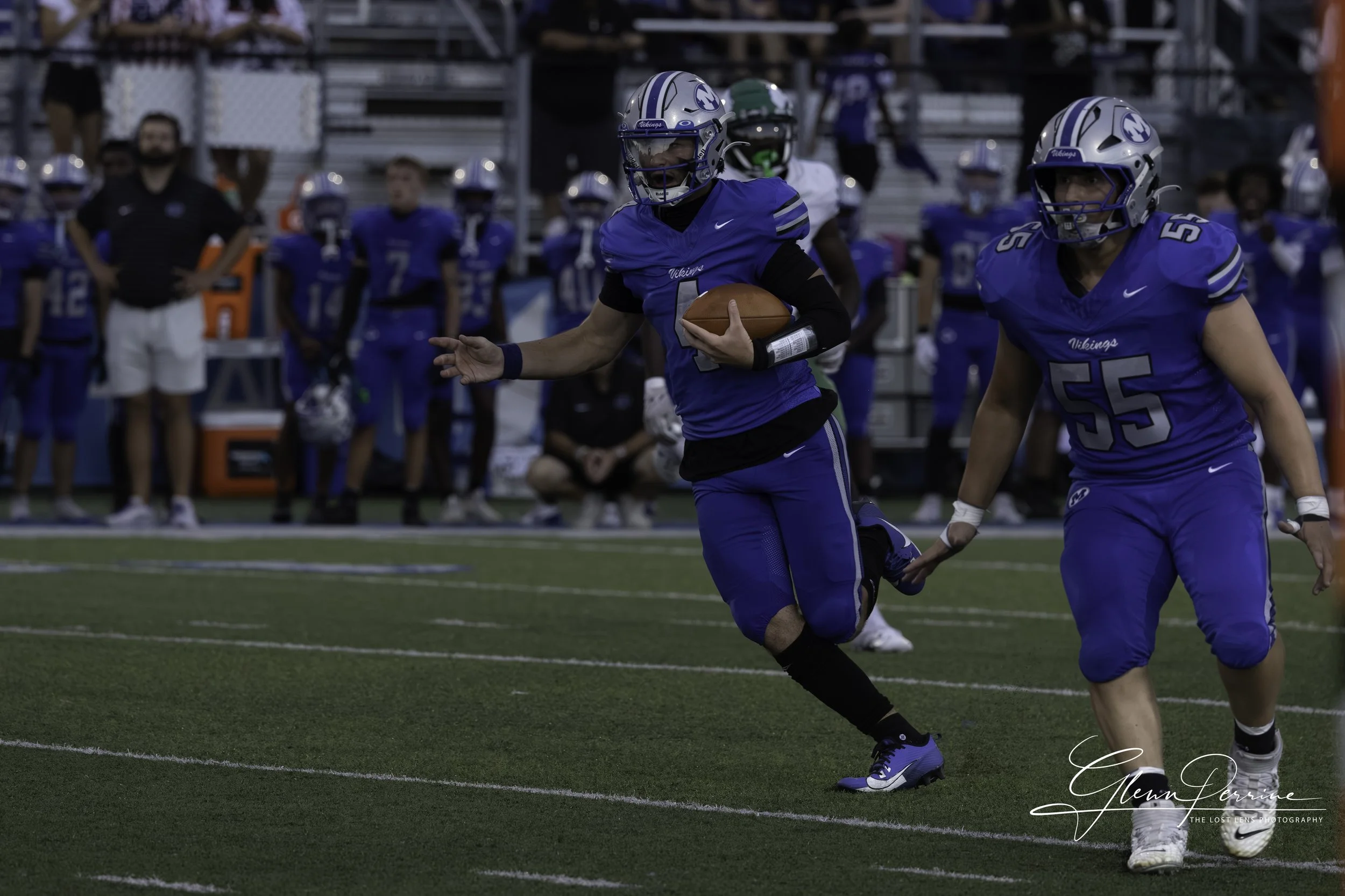 Football player in blue uniform holding a football running on the field with teammates and spectators in the background.