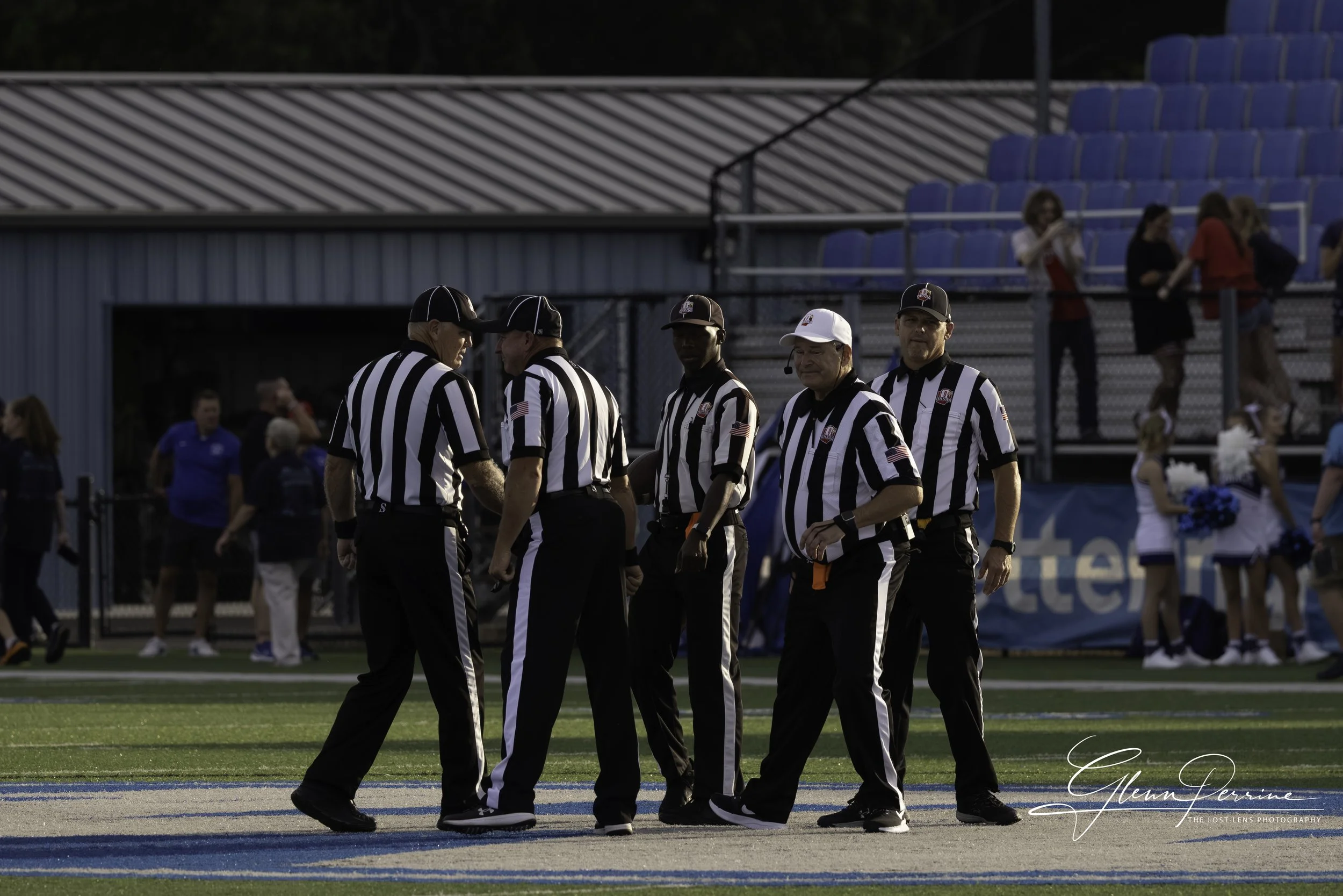 Six football referees standing on a football field during a game, with cheerleaders and spectators in the background.