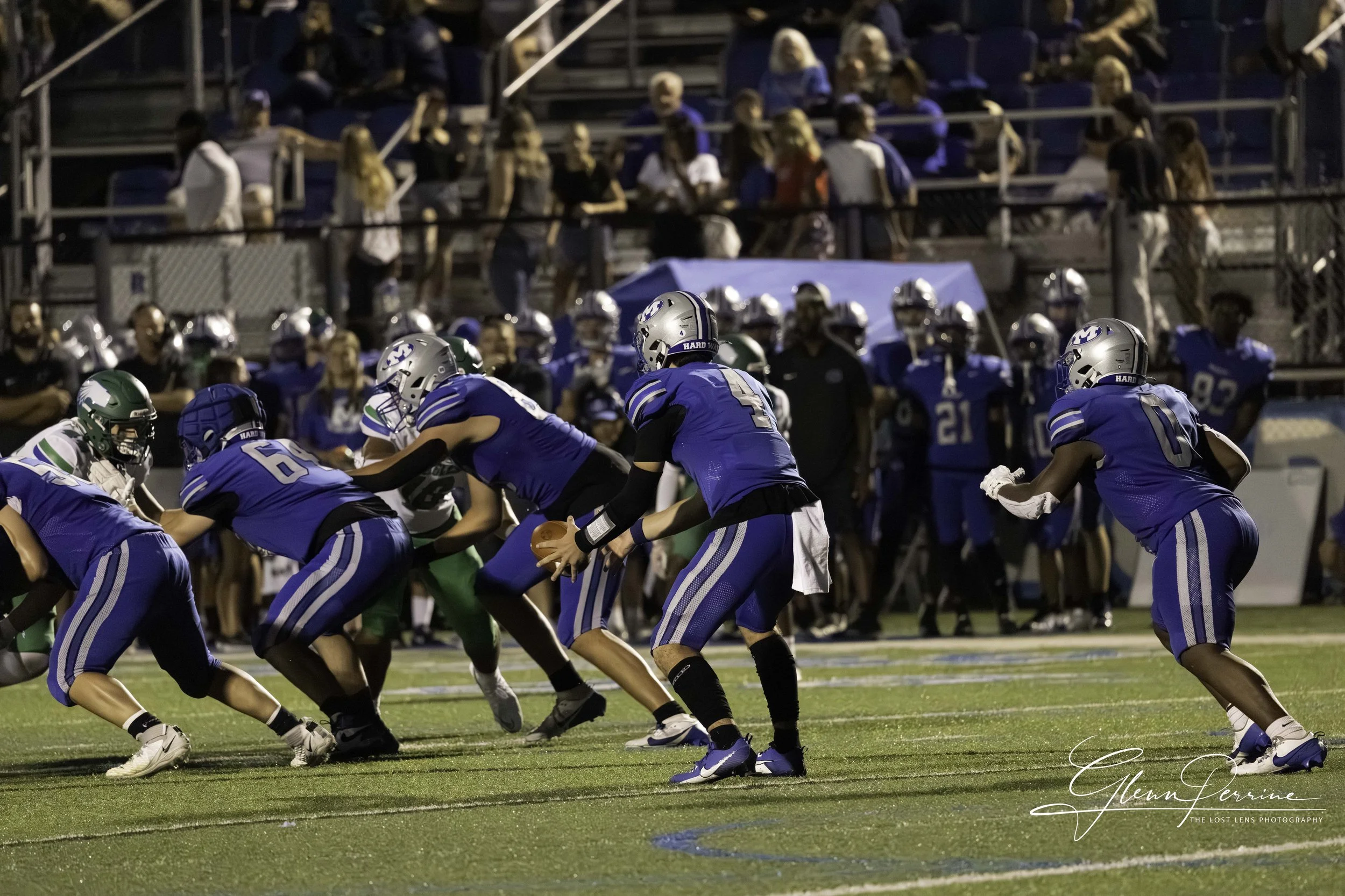 American football game at night with players in blue and green uniforms on the field, focusing on a blue team's quarterback about to pass the ball, with spectators in the background.
