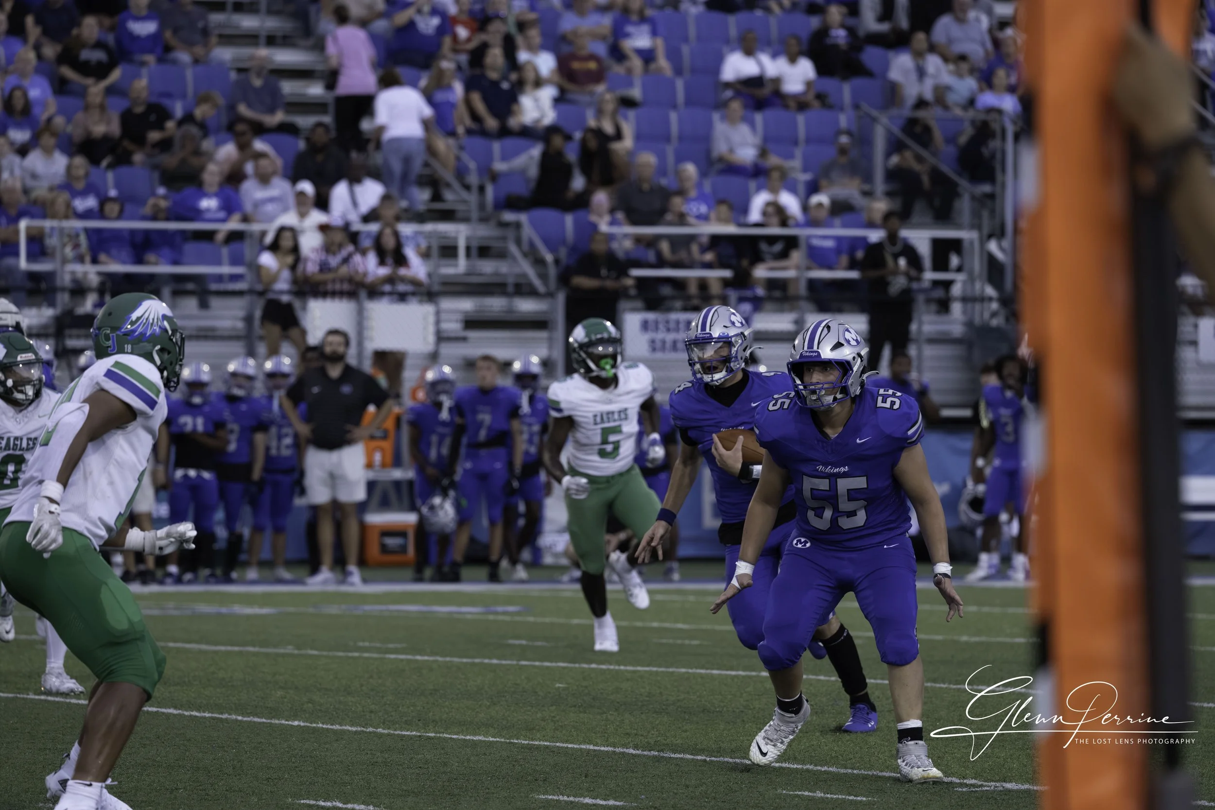 A football game with players on the field, one in blue uniform catching the ball, and others on both teams, some in green and white uniforms, in a stadium with spectators in the background.