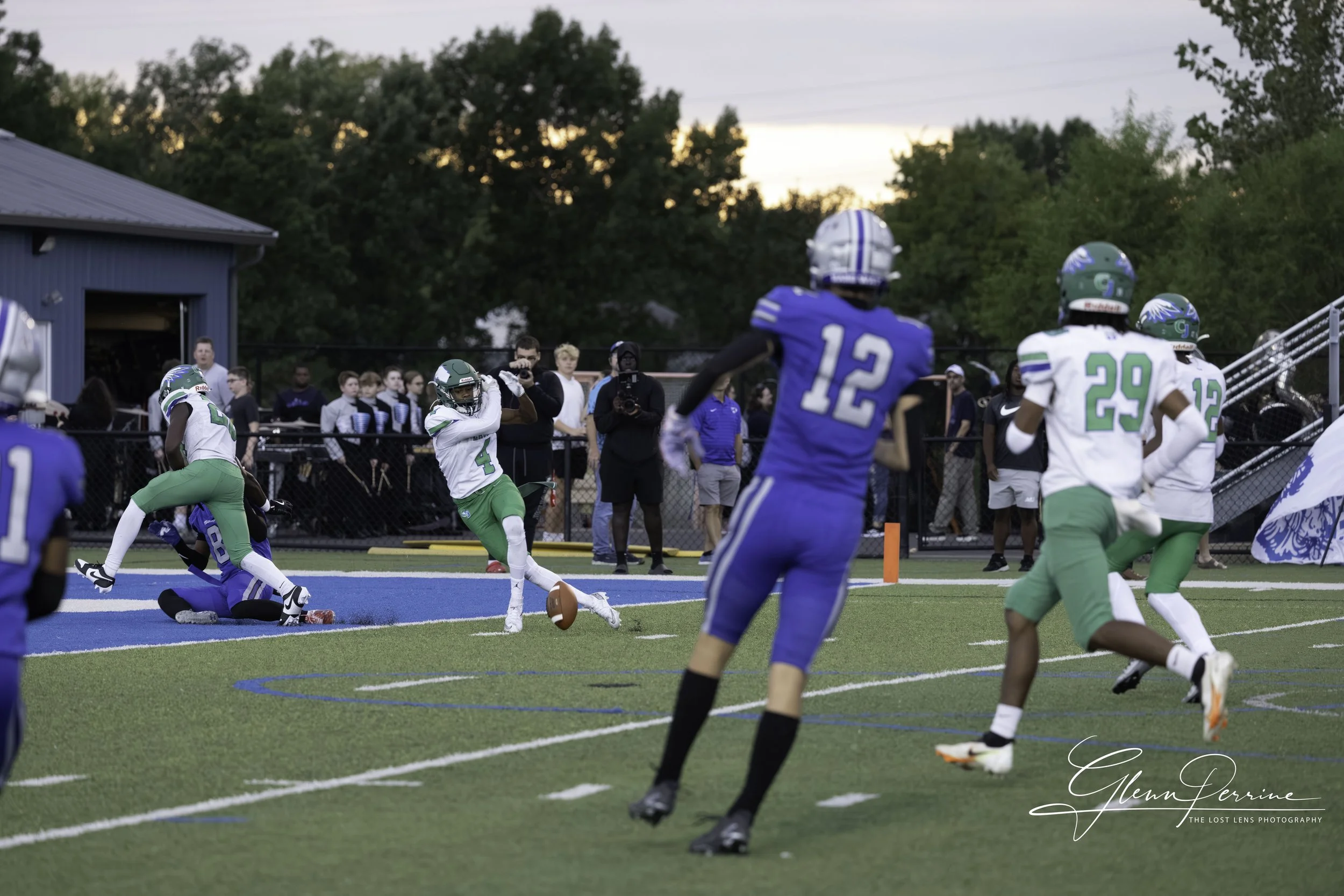 A youth football game with players in blue and green uniforms. A player in a white and green uniform is running with the football towards the end zone, while another green-uniformed player and a blue-uniformed player are on the ground. Several other 
