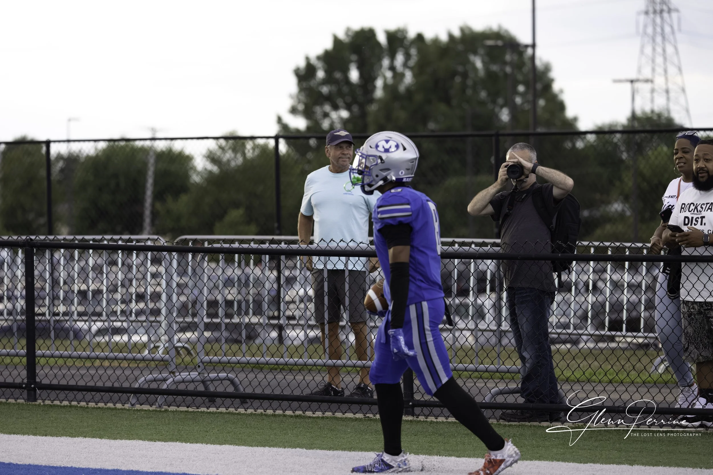 A football player in a blue uniform holding a football on a football field, with a fence and spectators watching in the background, including a man taking photos and a woman smiling.