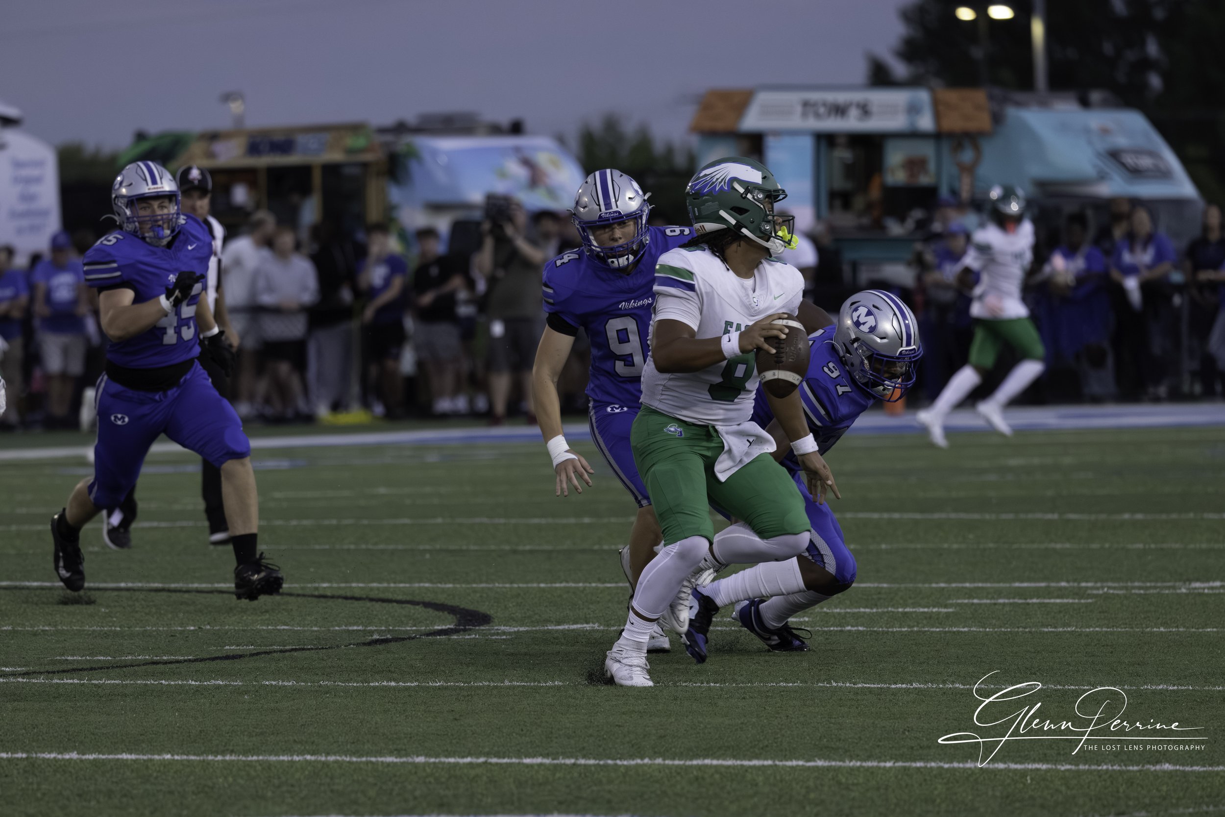 A football game with players in blue and white uniforms, with one player in a green uniform holding the ball and running.