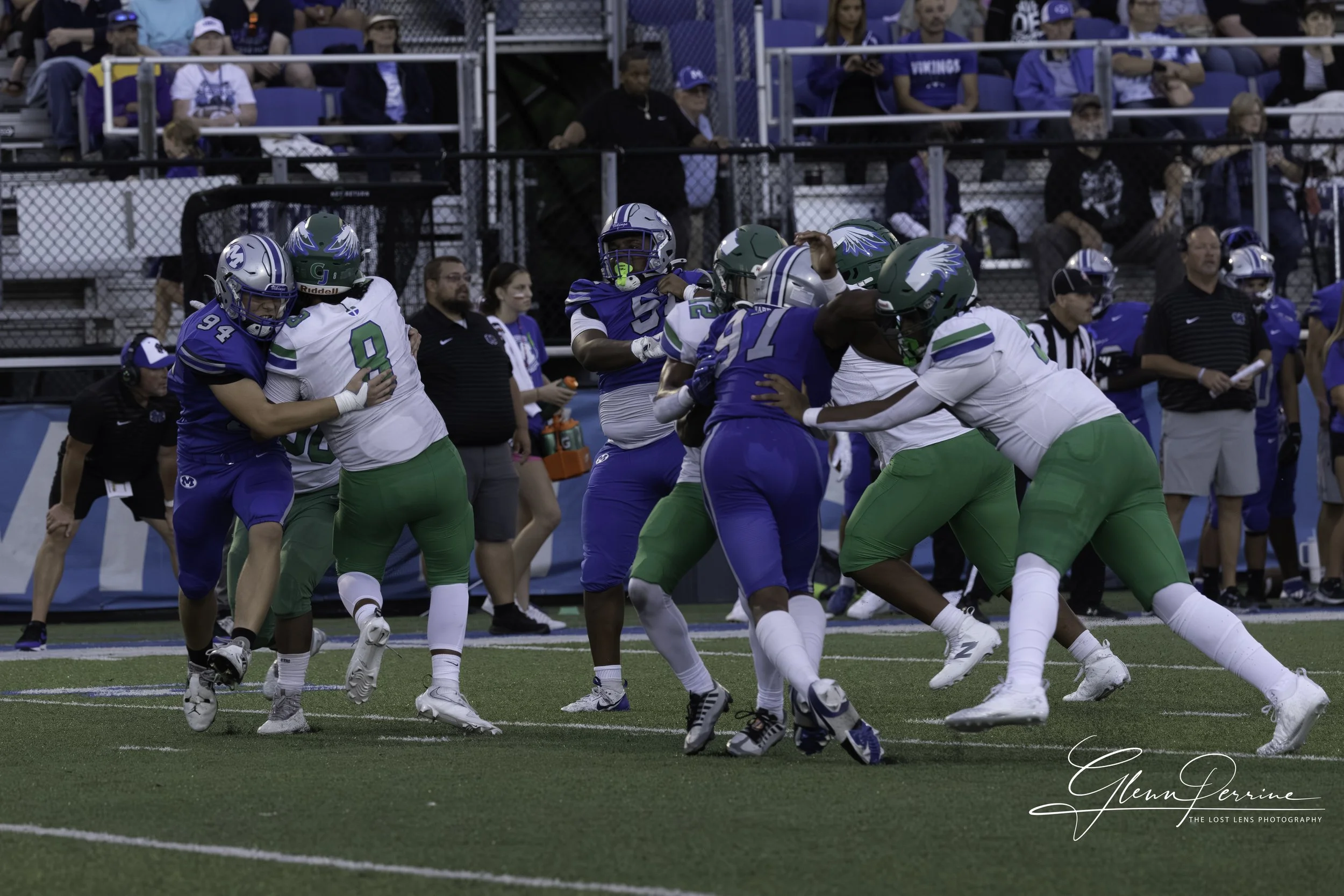 American football game with players from two teams, one in blue uniforms and the other in white and green, engaged in a tackle on the field, with spectators in the stands.