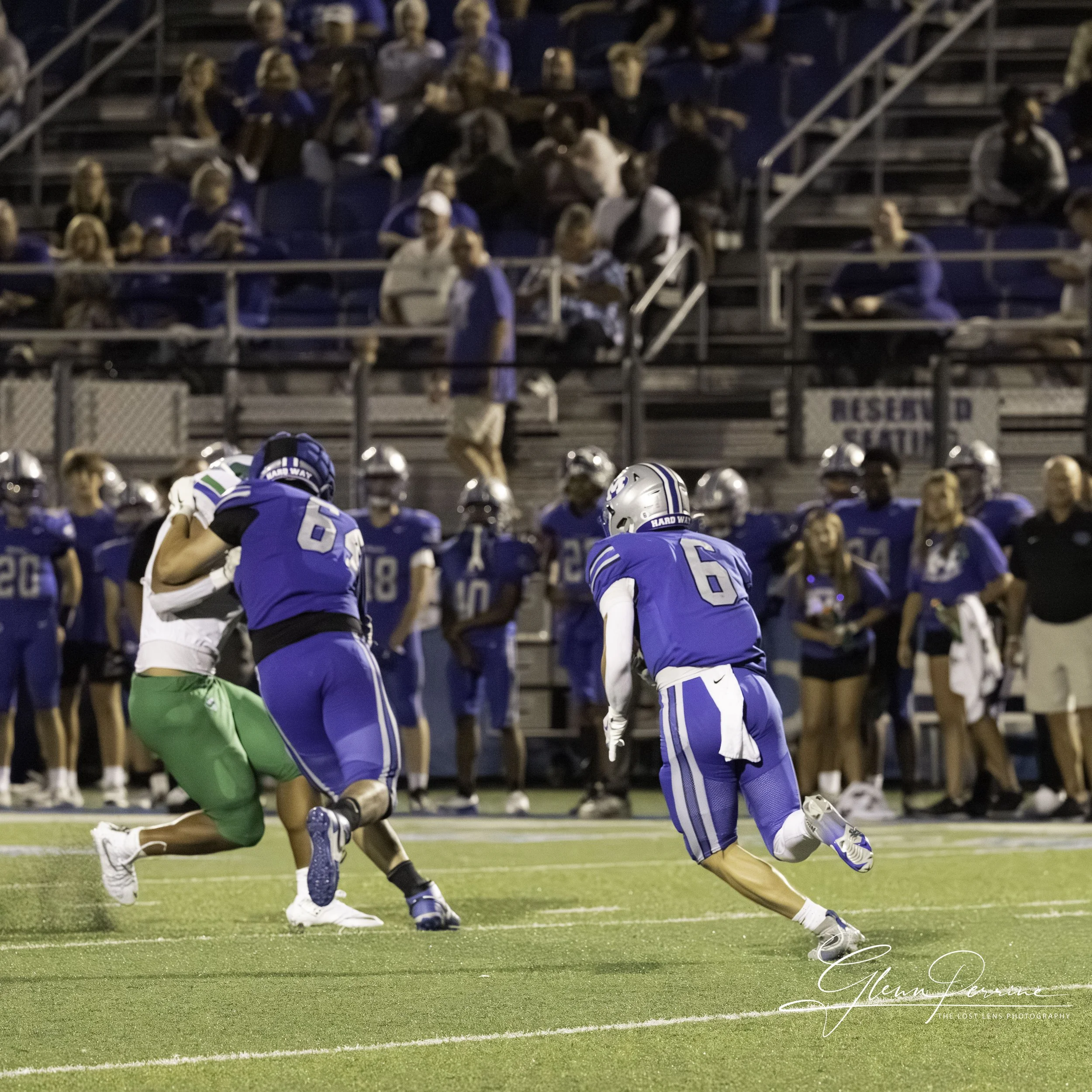 A football game at night showing players in blue and green uniforms, with two players in blue mid-play, one tackling a player in green, with spectators watching from bleachers in the background.