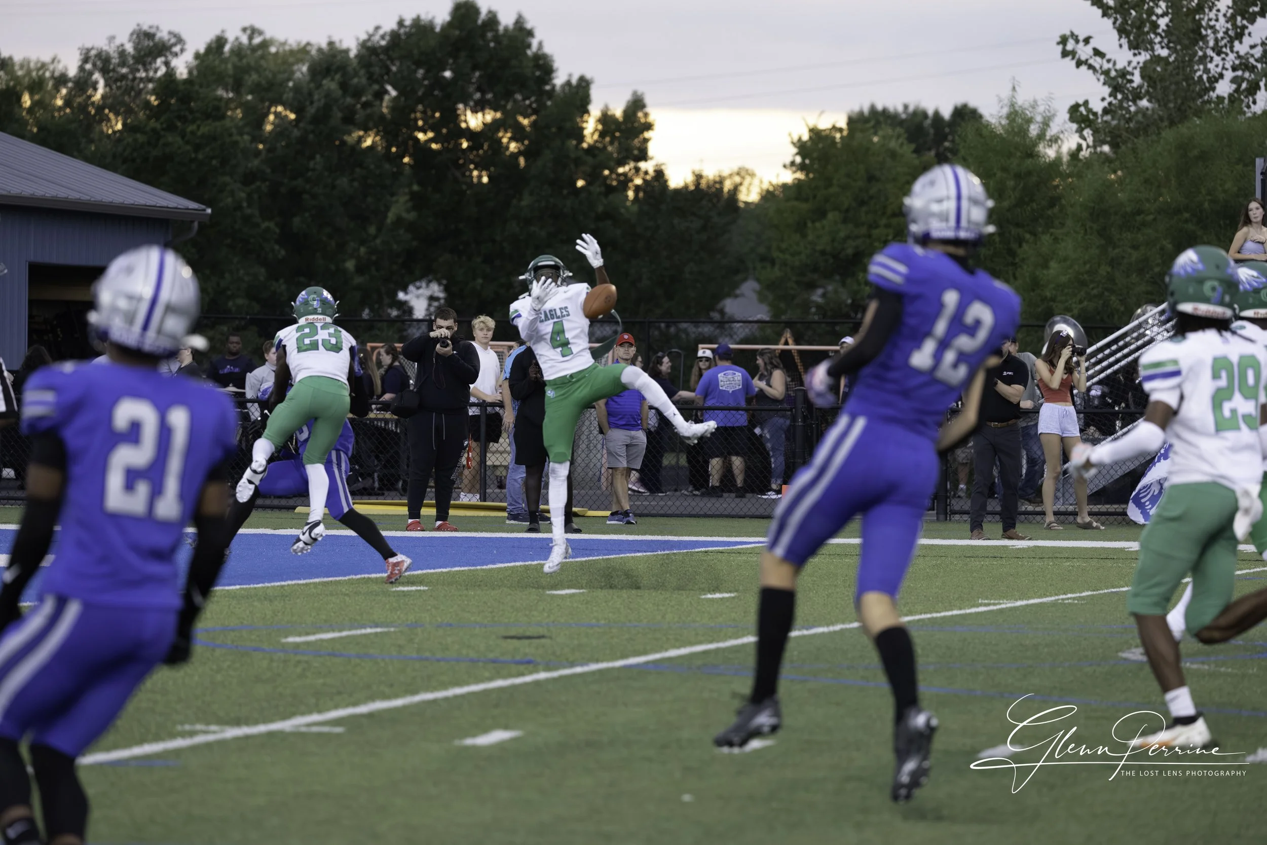 American football game in progress with players in blue and green uniforms on the field, some jumping and running while spectators watch from the sidelines during dusk.
