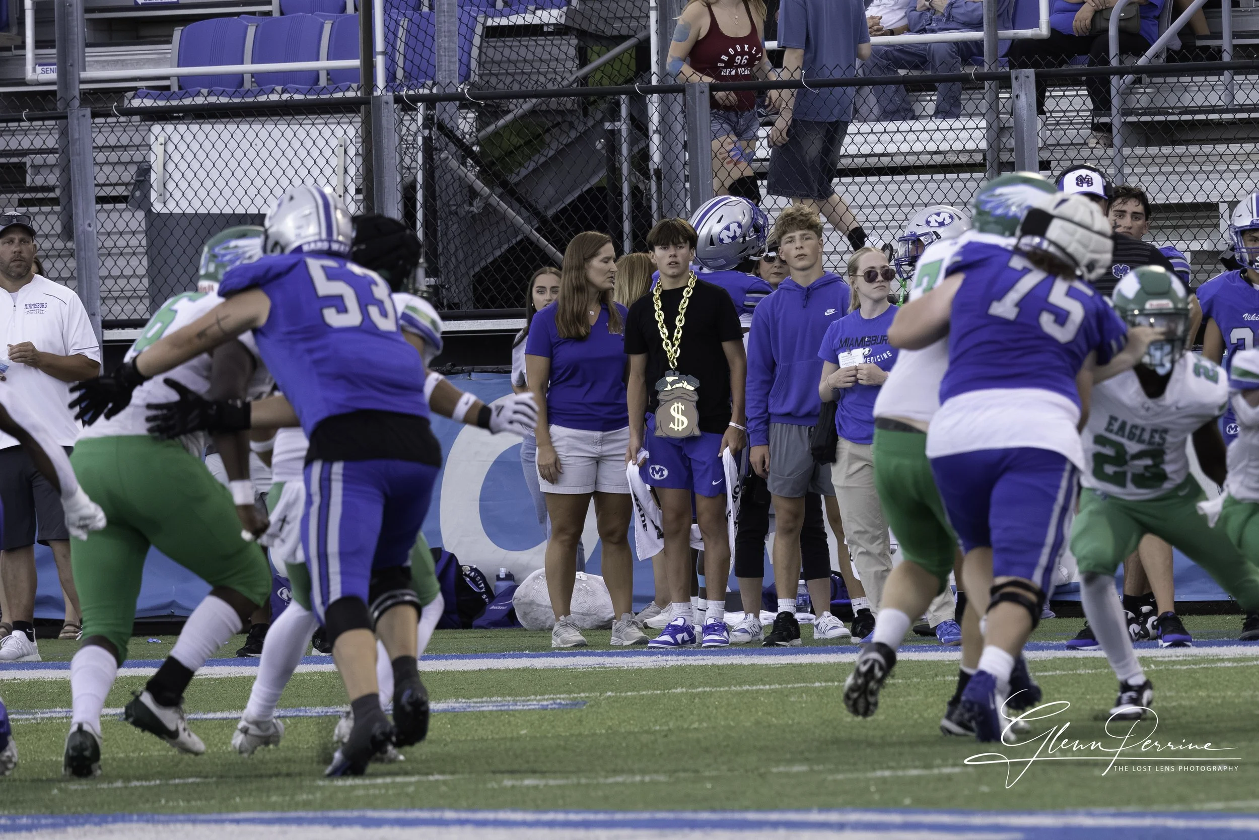 Panoramic view of a college football game with players in blue and green uniforms on the field. Spectators are watching from the bleachers and standing behind a chain-link fence.