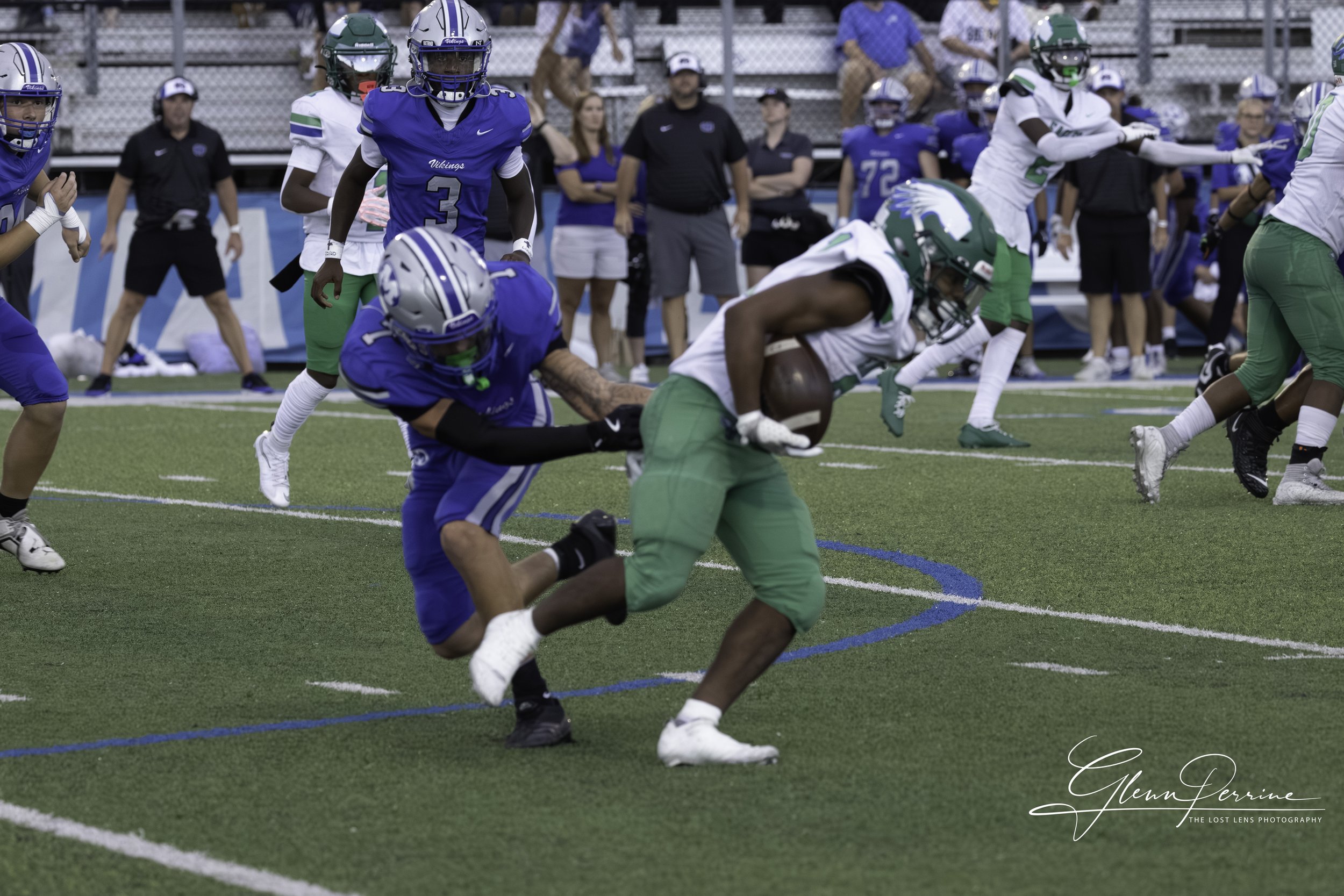 An American football game with players in blue and green uniforms. A player in green is running with the football while a player in blue tries to tackle him from behind. Spectators watch from the bleachers in the background.
