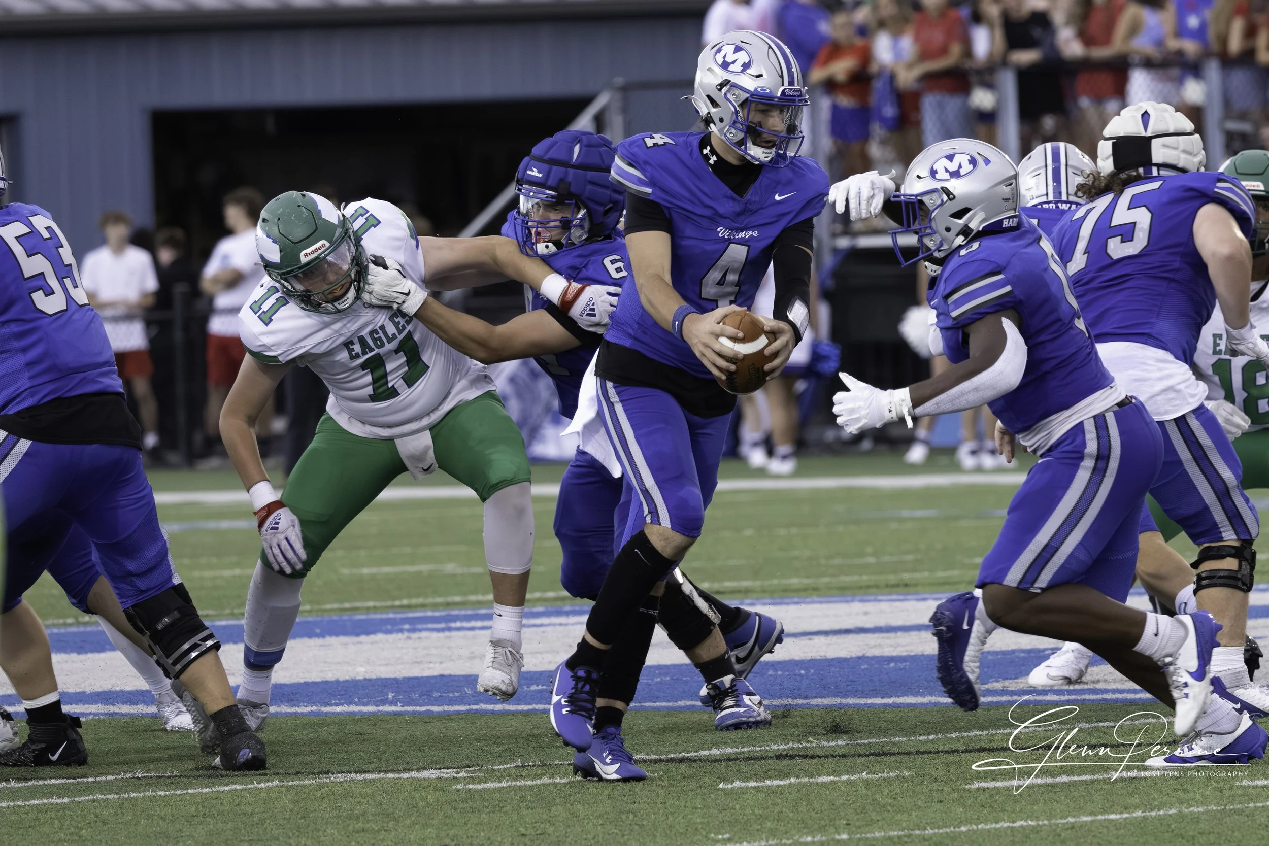 An American football game in progress with a player in a blue uniform holding the ball, surrounded by opponents and teammates on the field.