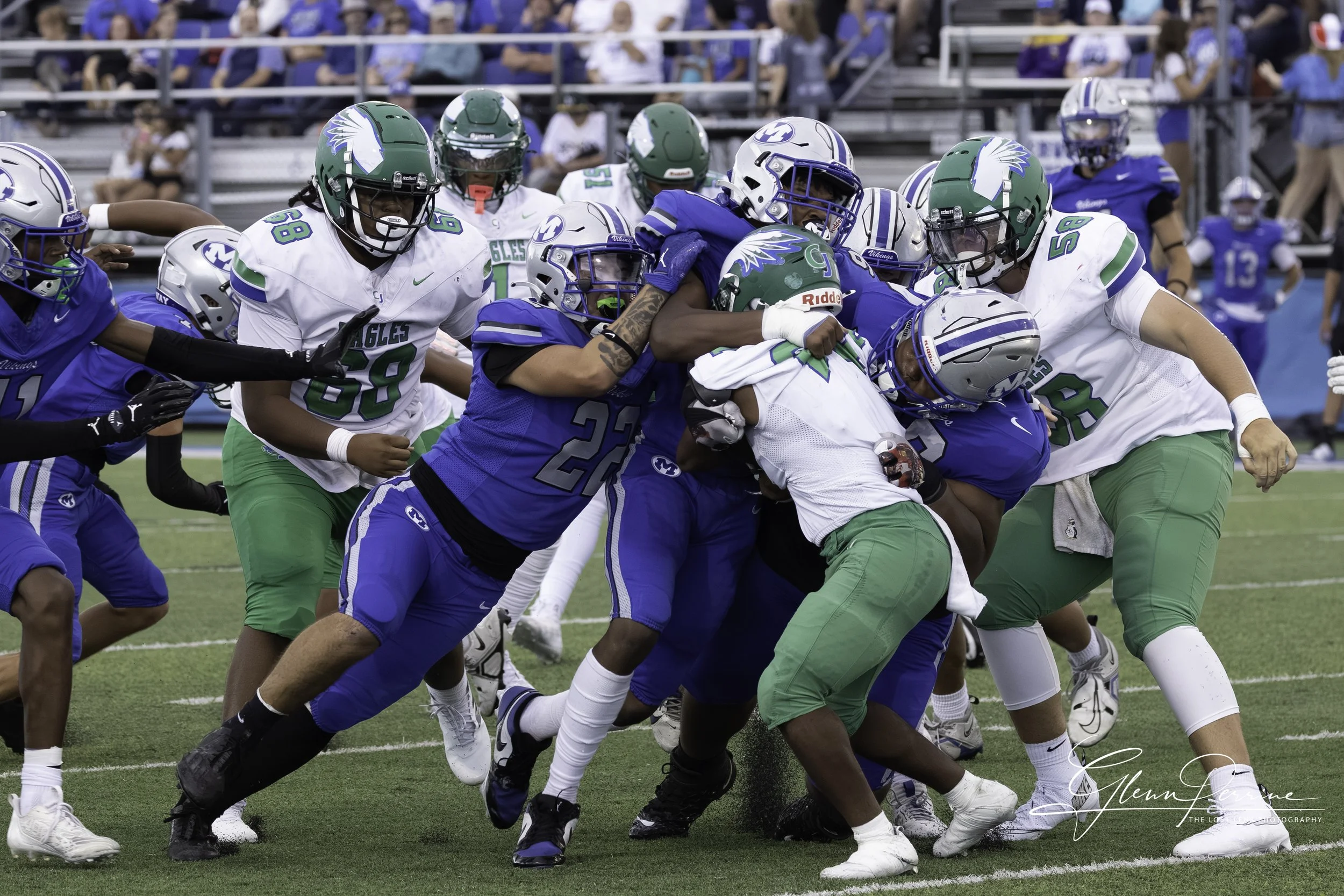 A football game with players from two teams, one in green and white uniforms and the other in blue uniforms, engaged in a tackle on the field.