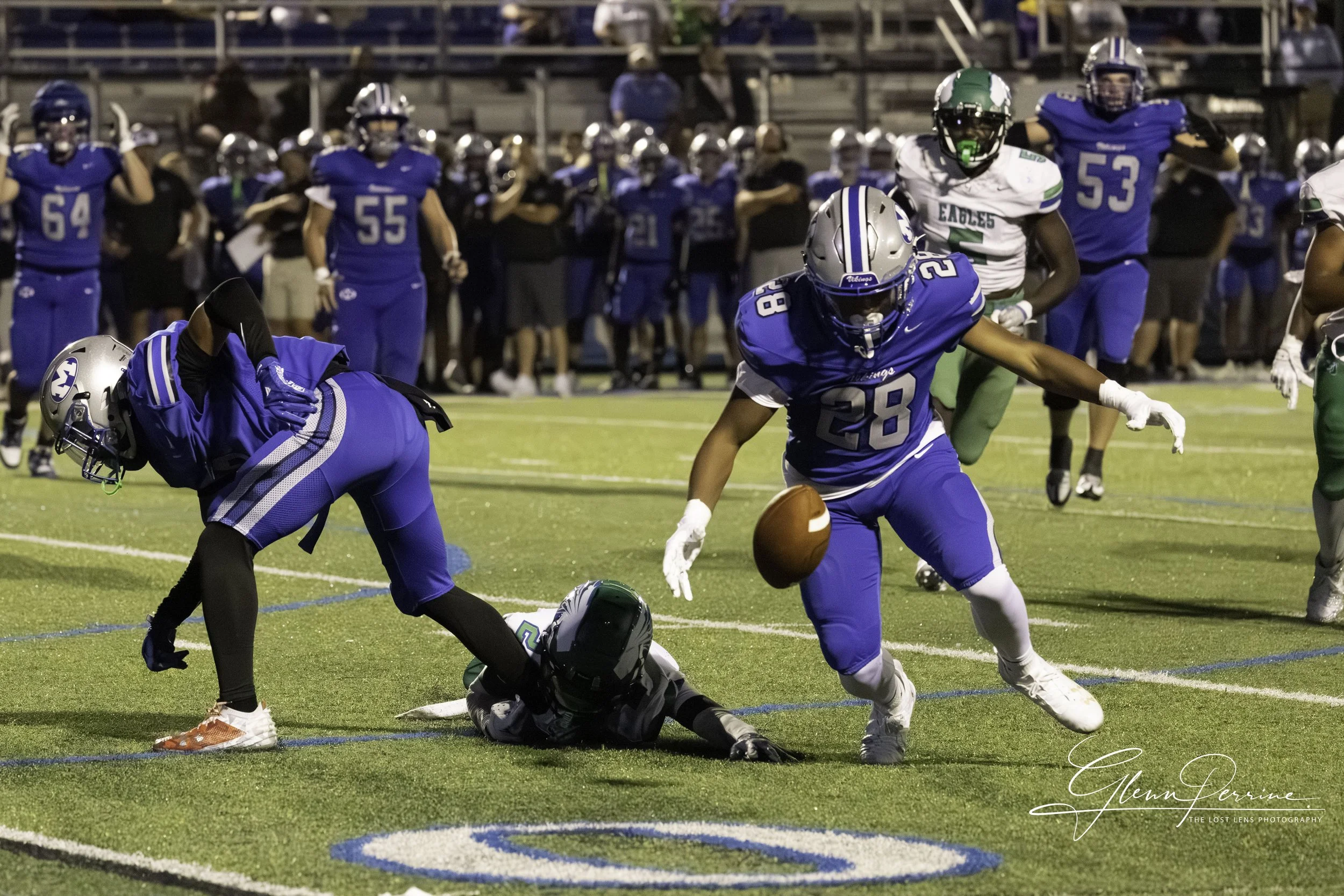 Football players in blue and green uniforms during a game, with one blue player reaching for a ball while another player is on the ground, and a crowd in the background.