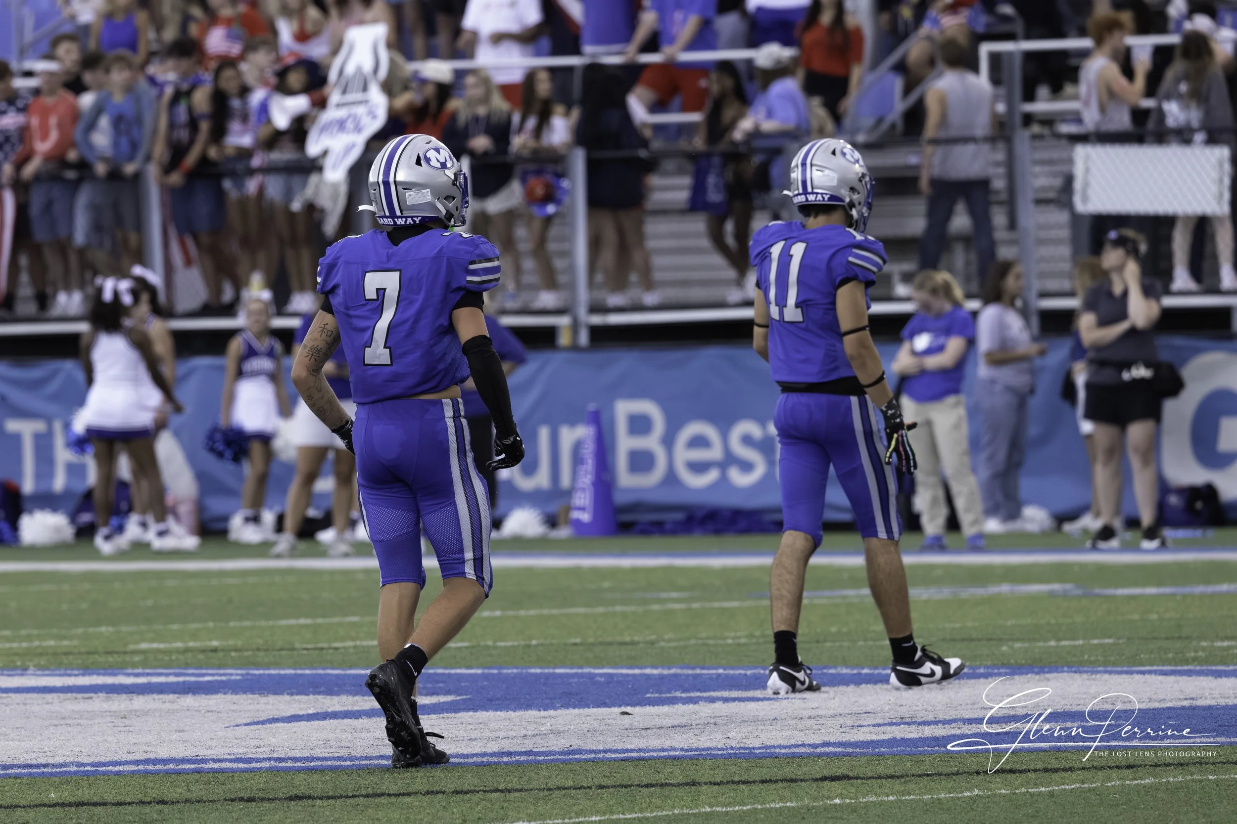Two American football players in blue uniforms and helmets standing on the field during a game, with spectators in the background.