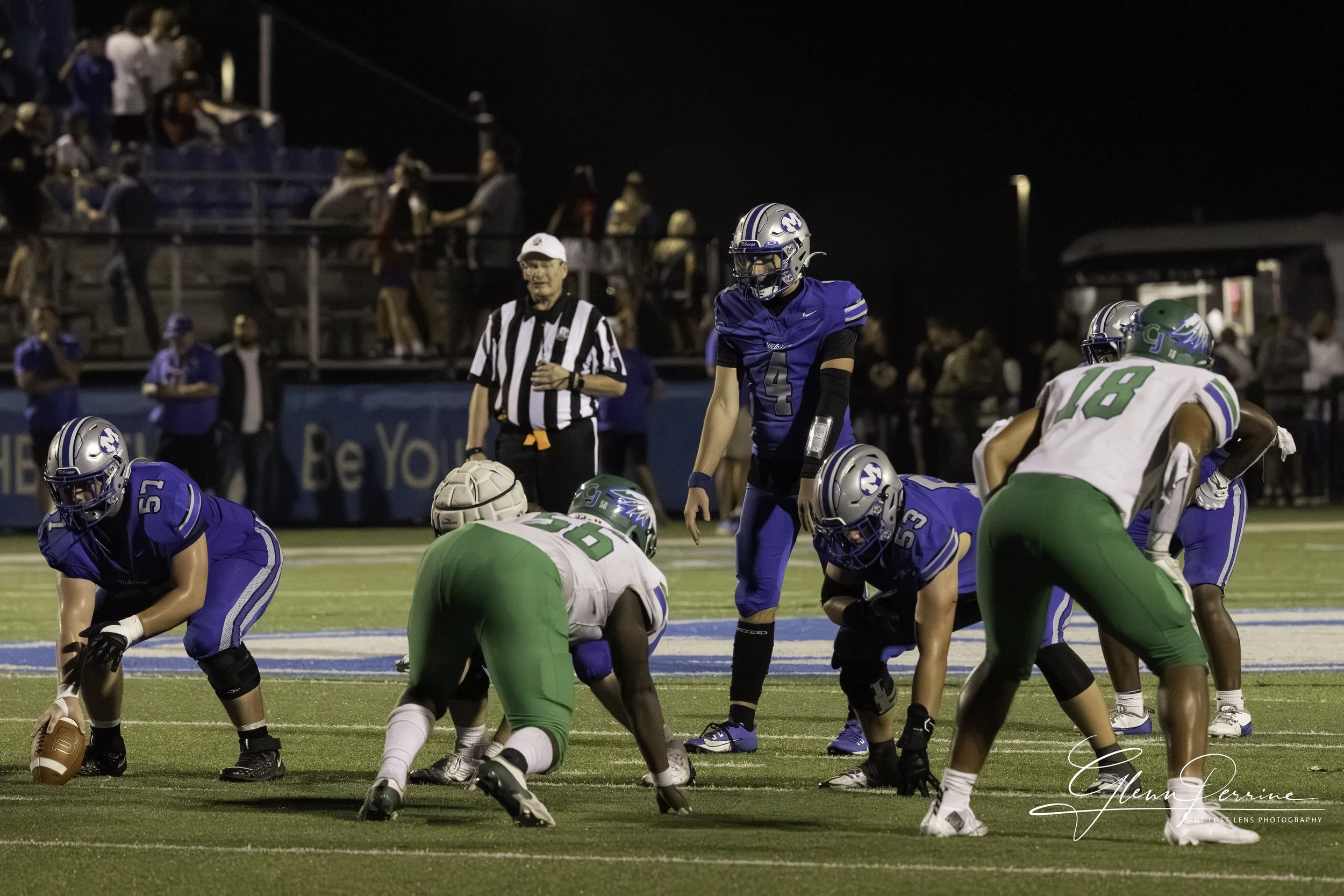 A nighttime American football game with players in blue and green uniforms on the field, a referee, and spectators in the background.
