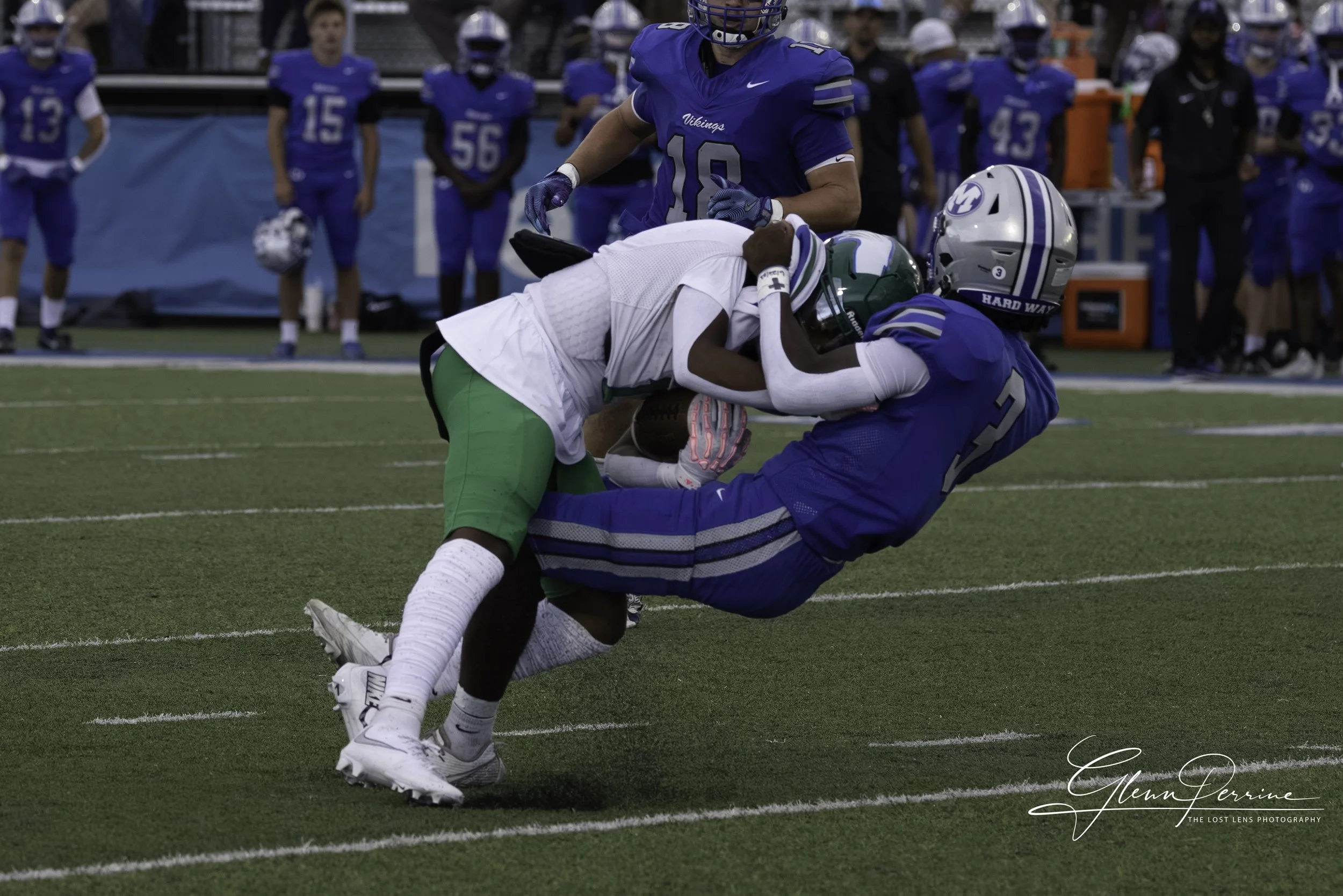 Two football players from opposing teams tackle each other on the field, with other players and coaches on the sideline in the background.