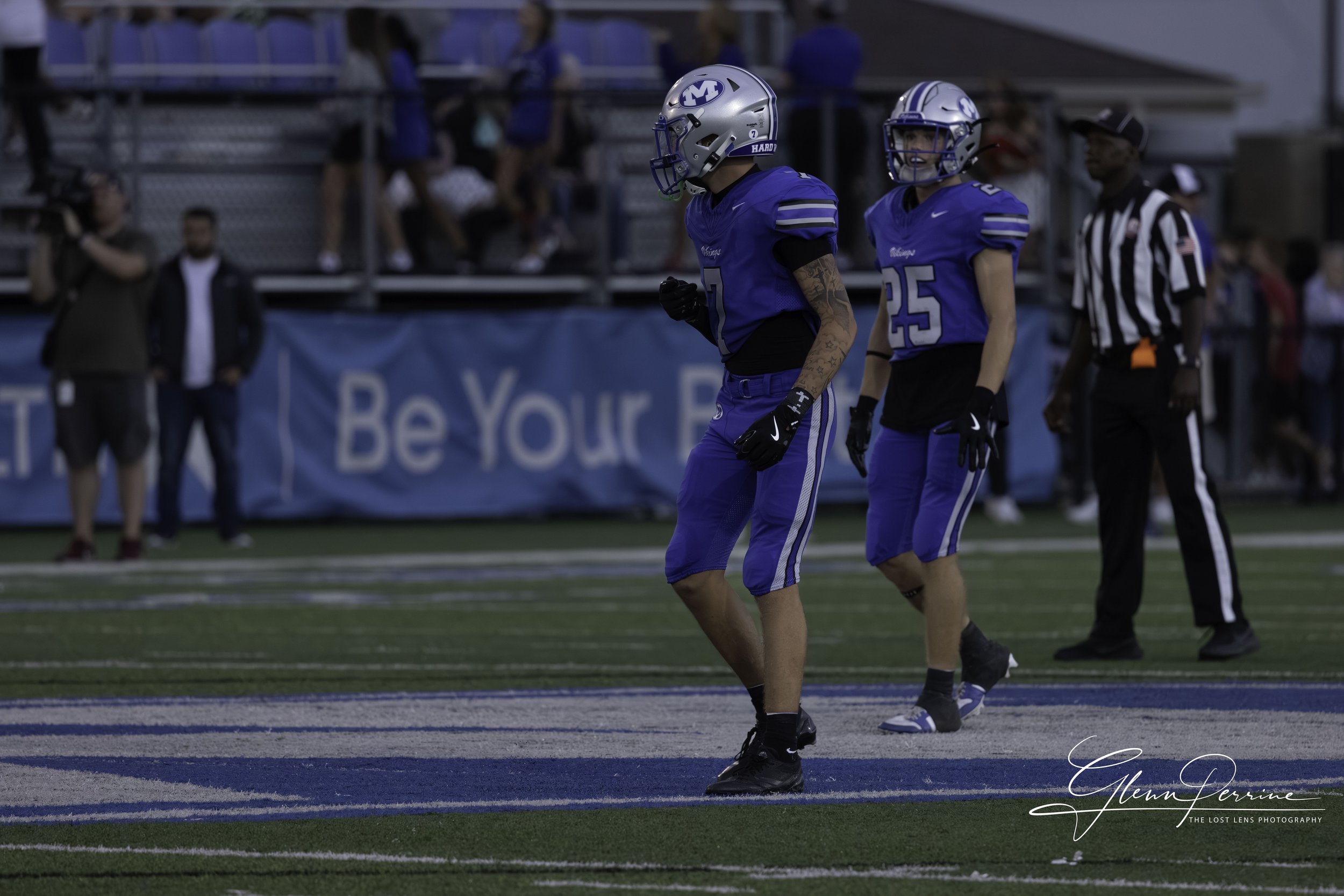 Two football players in blue jerseys and helmets standing on a football field during a game, with a referee and spectators in the background.