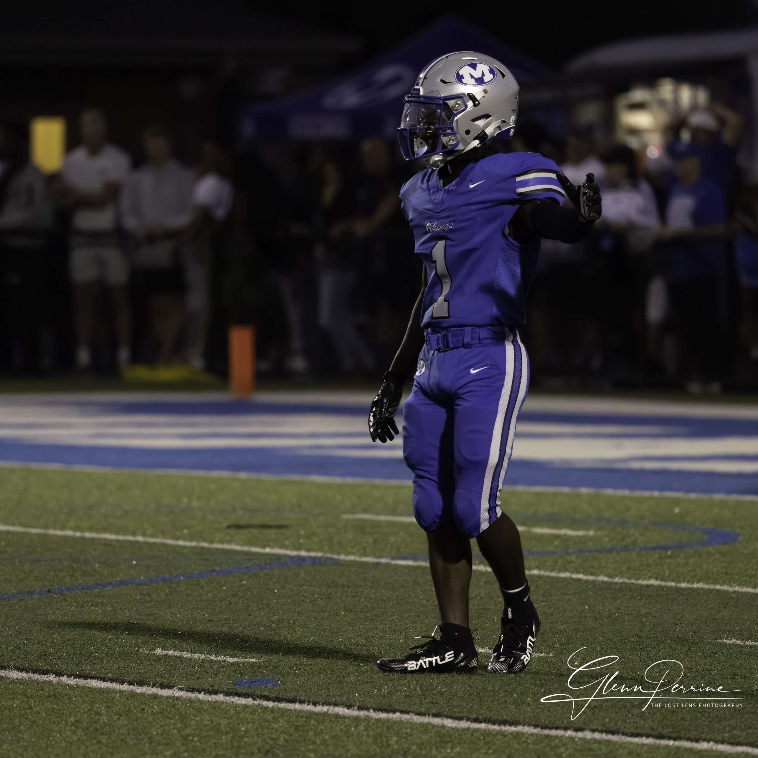 A football player in a blue uniform with a helmet stands on a football field at night, with spectators in the background.