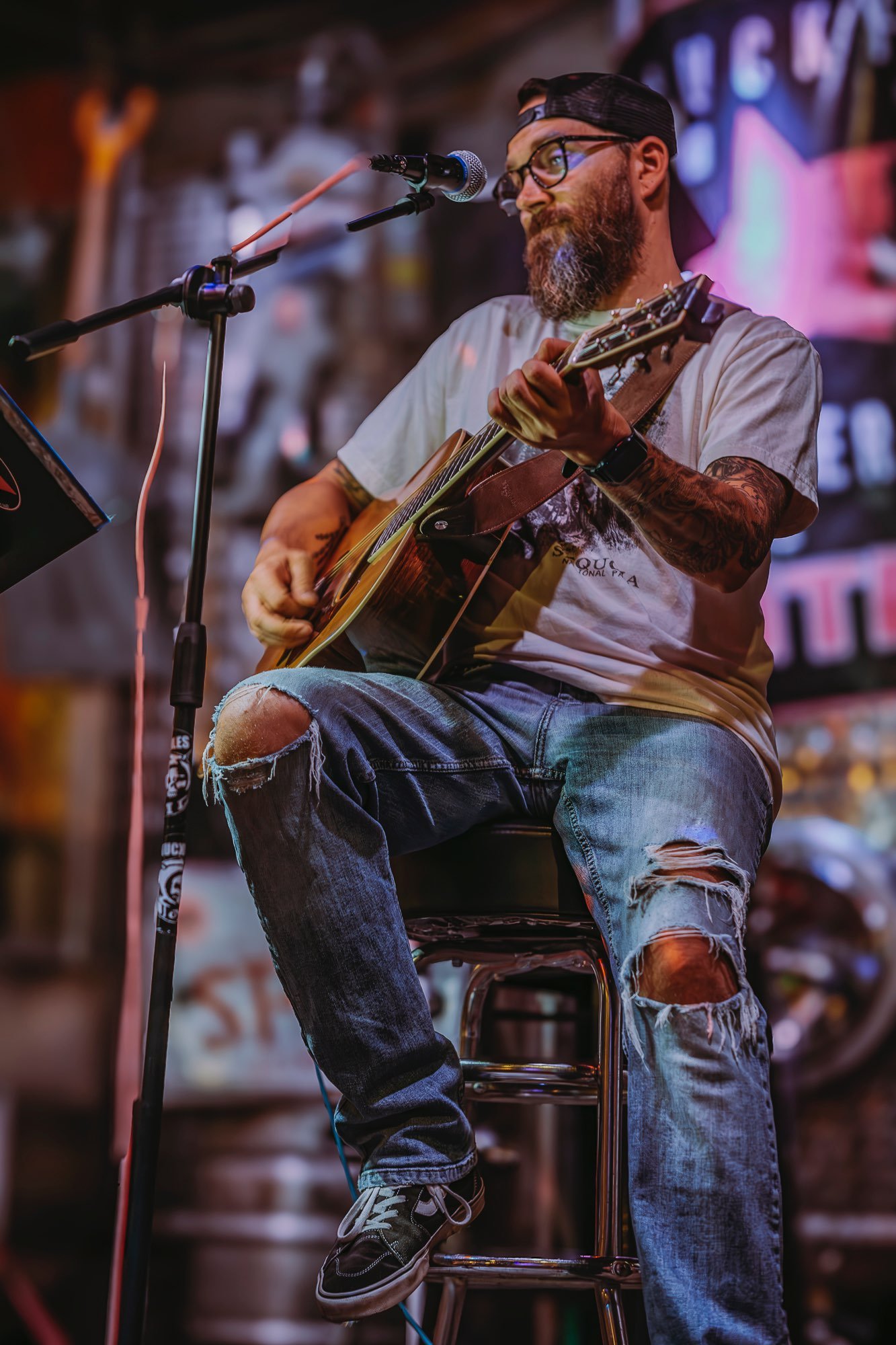 A man with tattoos, glasses, a beard, and a backwards cap playing an acoustic guitar on stage, with a microphone in front of him.