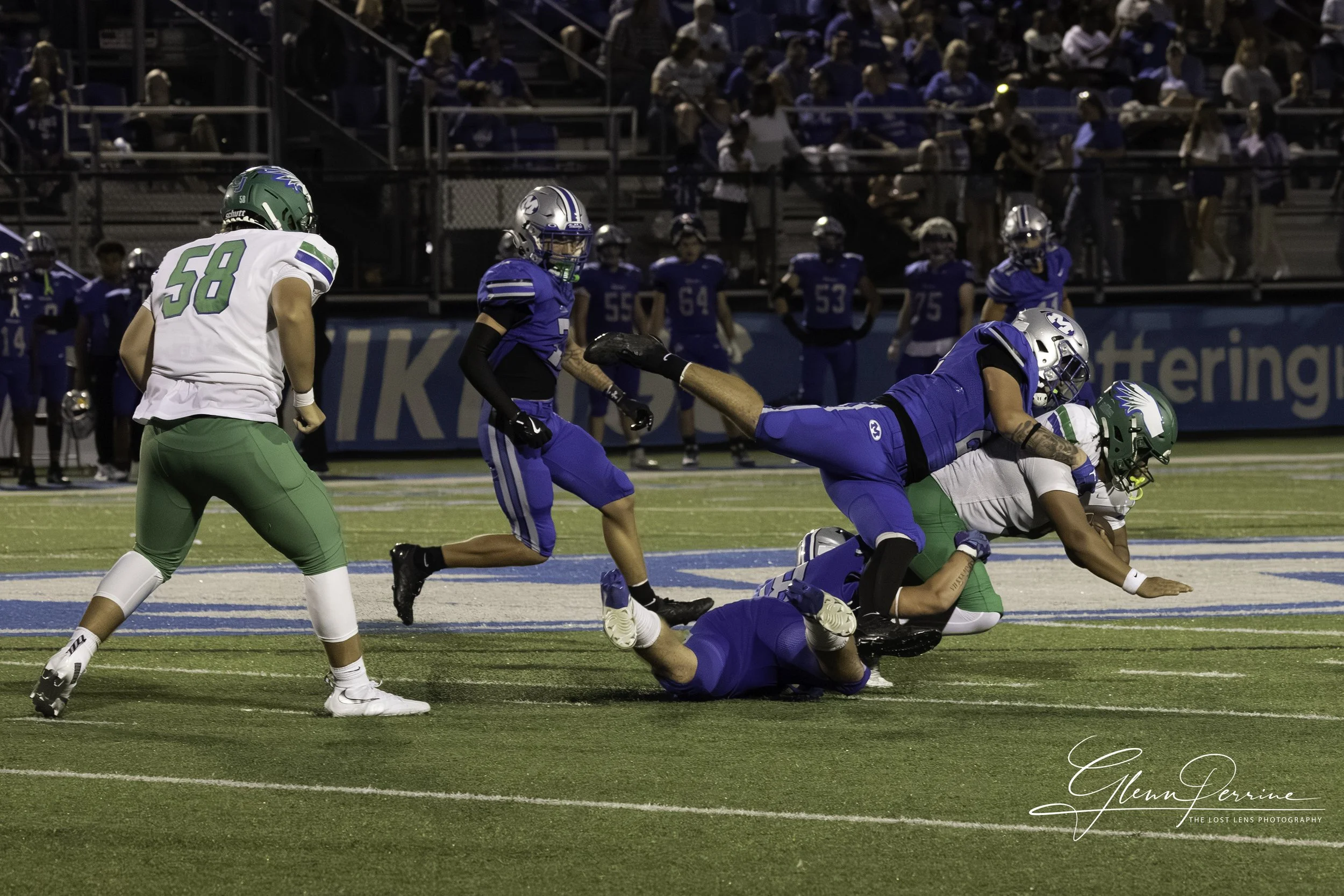 An American football game in progress with players tackling on the field; players are dressed in blue and white uniforms, with the crowd watching from the stands in the background.