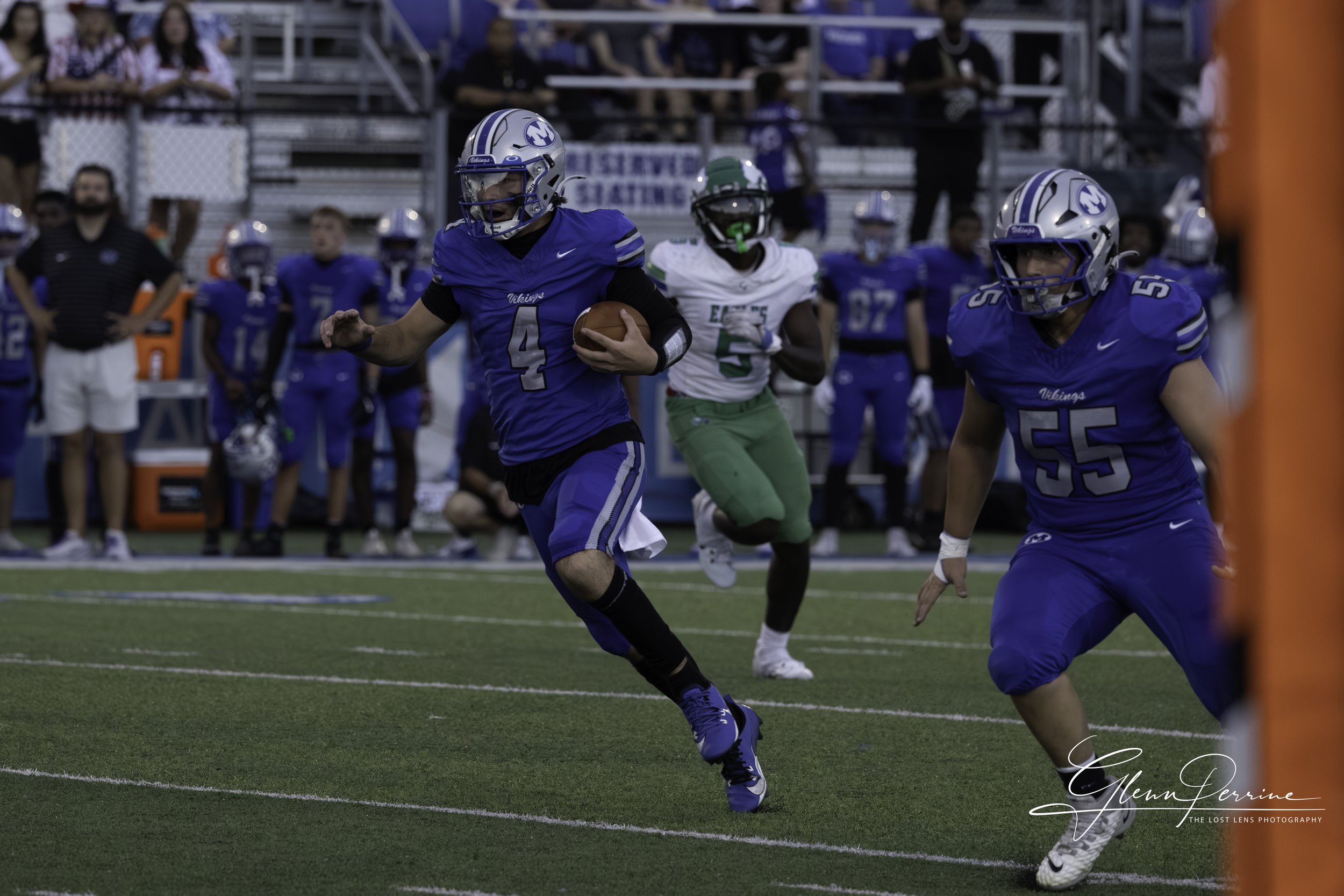 A football player in a blue uniform running with the ball on the field while other players and spectators watch from the sidelines.