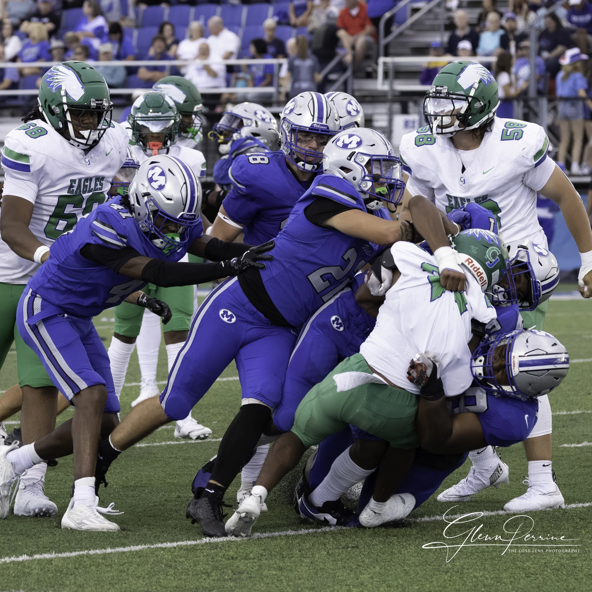 American football players in blue and white uniforms competing for the ball on the field, with spectators in the background.