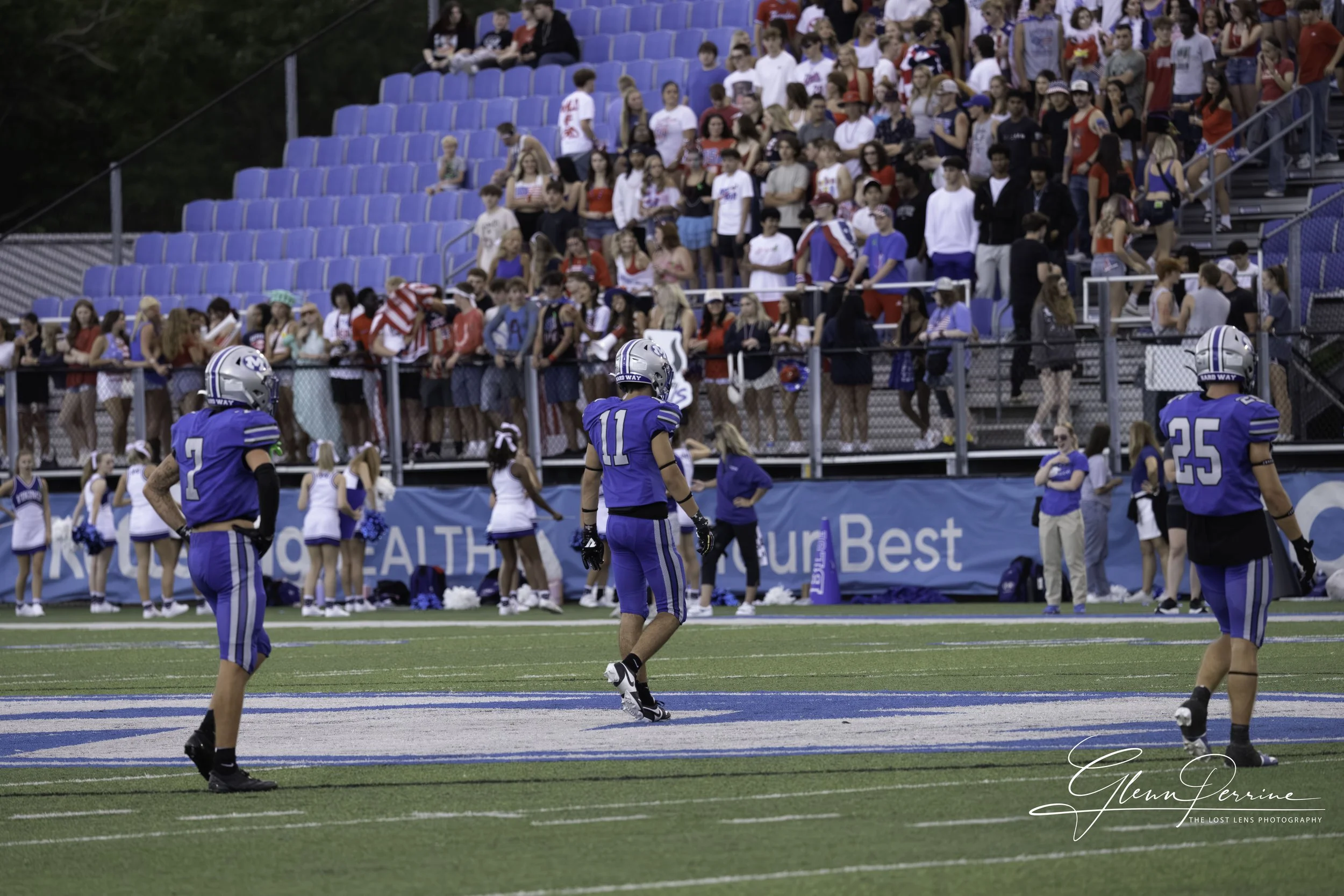 Three football players in blue uniforms and helmets stand on a football field during a game, with a crowd of spectators and cheerleaders in the background.