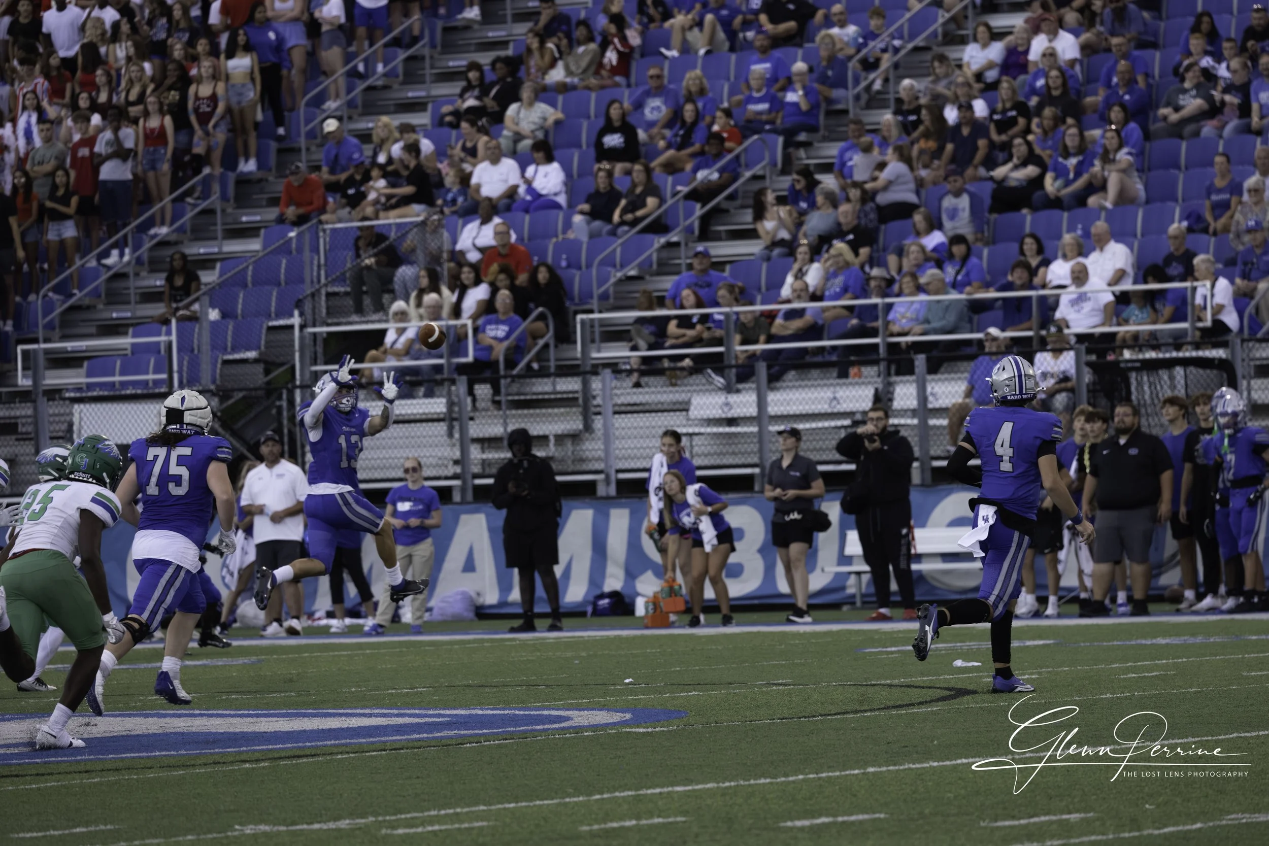 A football game with a player in blue uniform catching a football in the end zone with players and crowd watching.