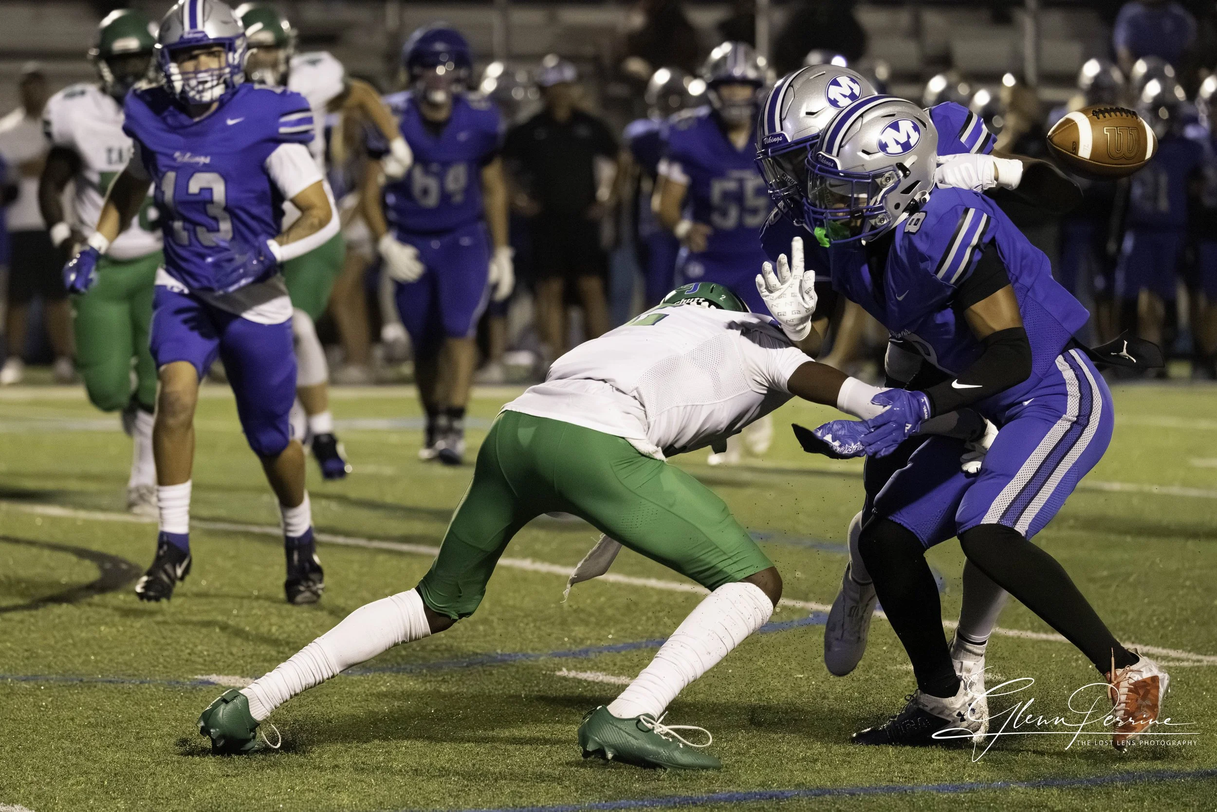 A football game with players in blue and white uniforms. One blue player is tackling a white and green player trying to catch the football. Other players and spectators are visible in the background.
