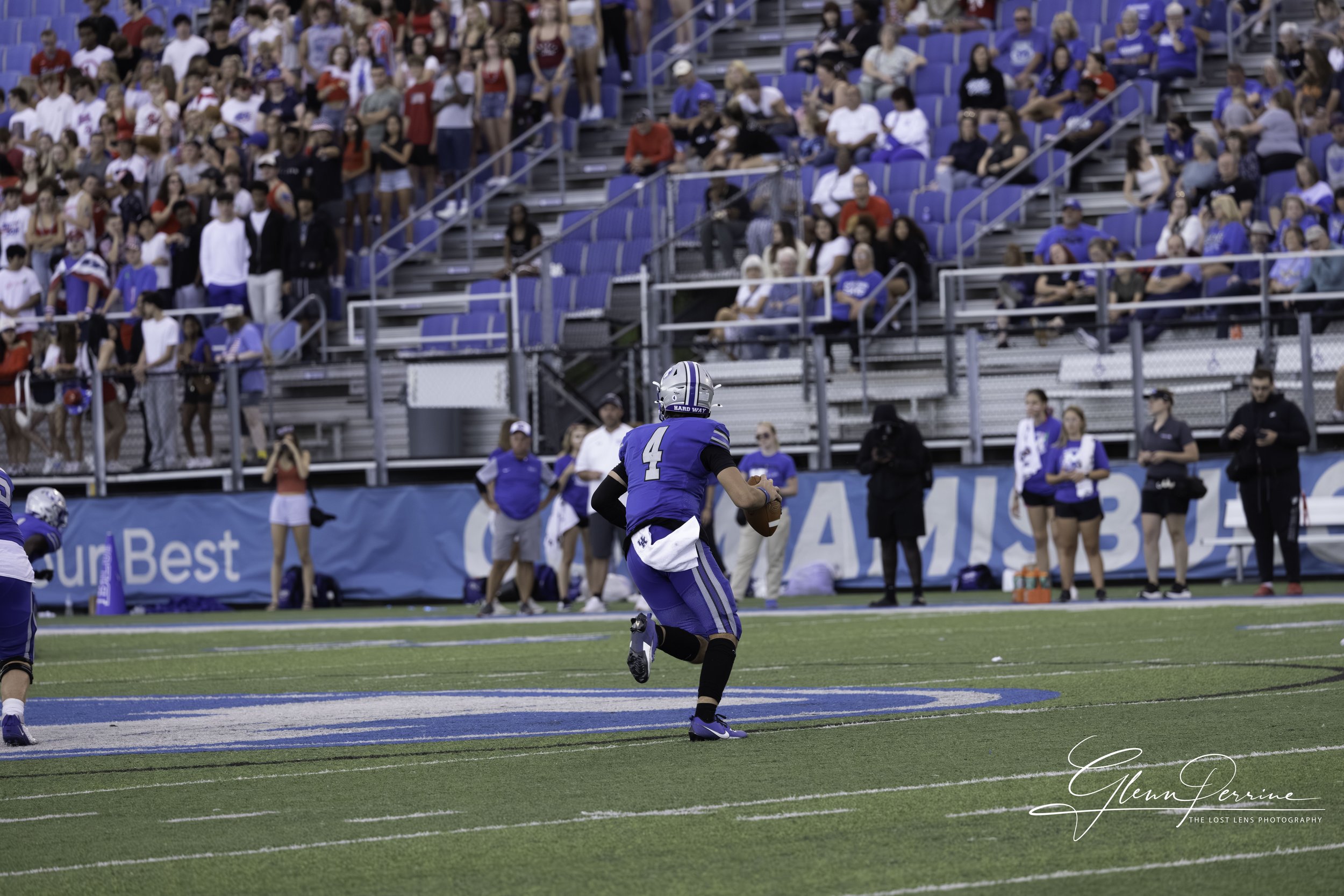 A football player wearing a blue uniform with the number 4 is running on the field holding a football. The stadium is filled with spectators, many of whom are standing and watching the game.