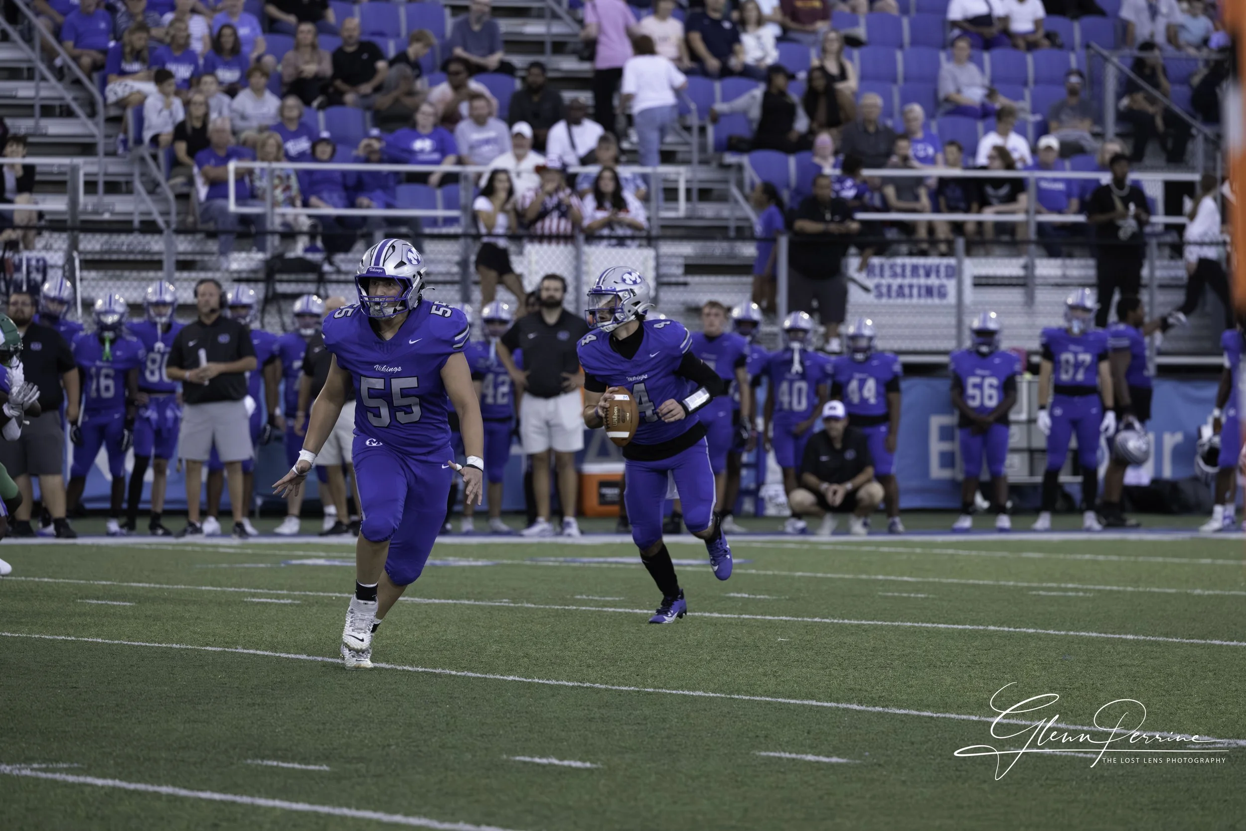 A football game in progress with players in blue and black uniforms on the field, a player holding the football running, and spectators watching from the bleachers.