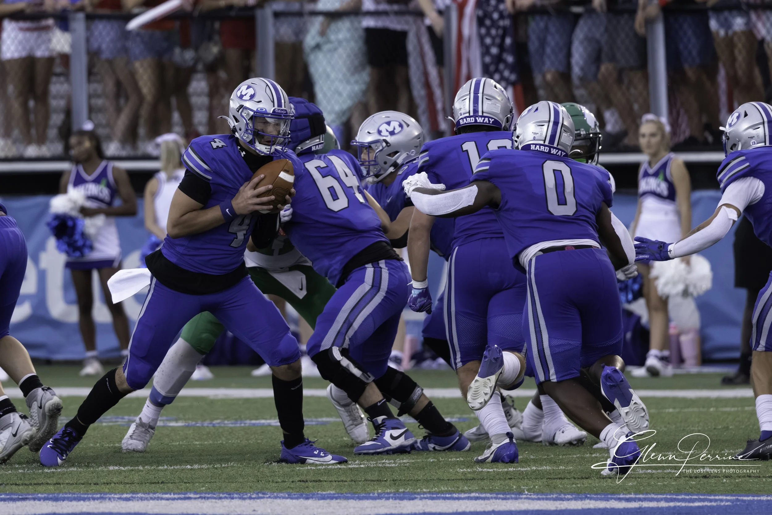 American football game with players in blue uniforms, one holding the ball, others blocking on the field with cheerleaders and spectators in the background.