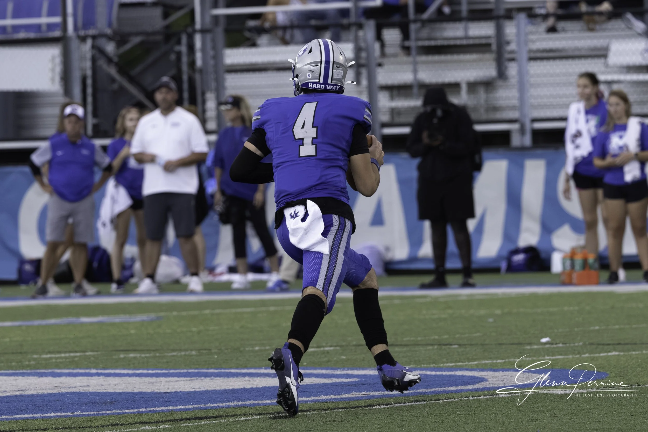 A football player in blue uniform running on the field with a helmet, with teammates and coaches watching from the sidelines.