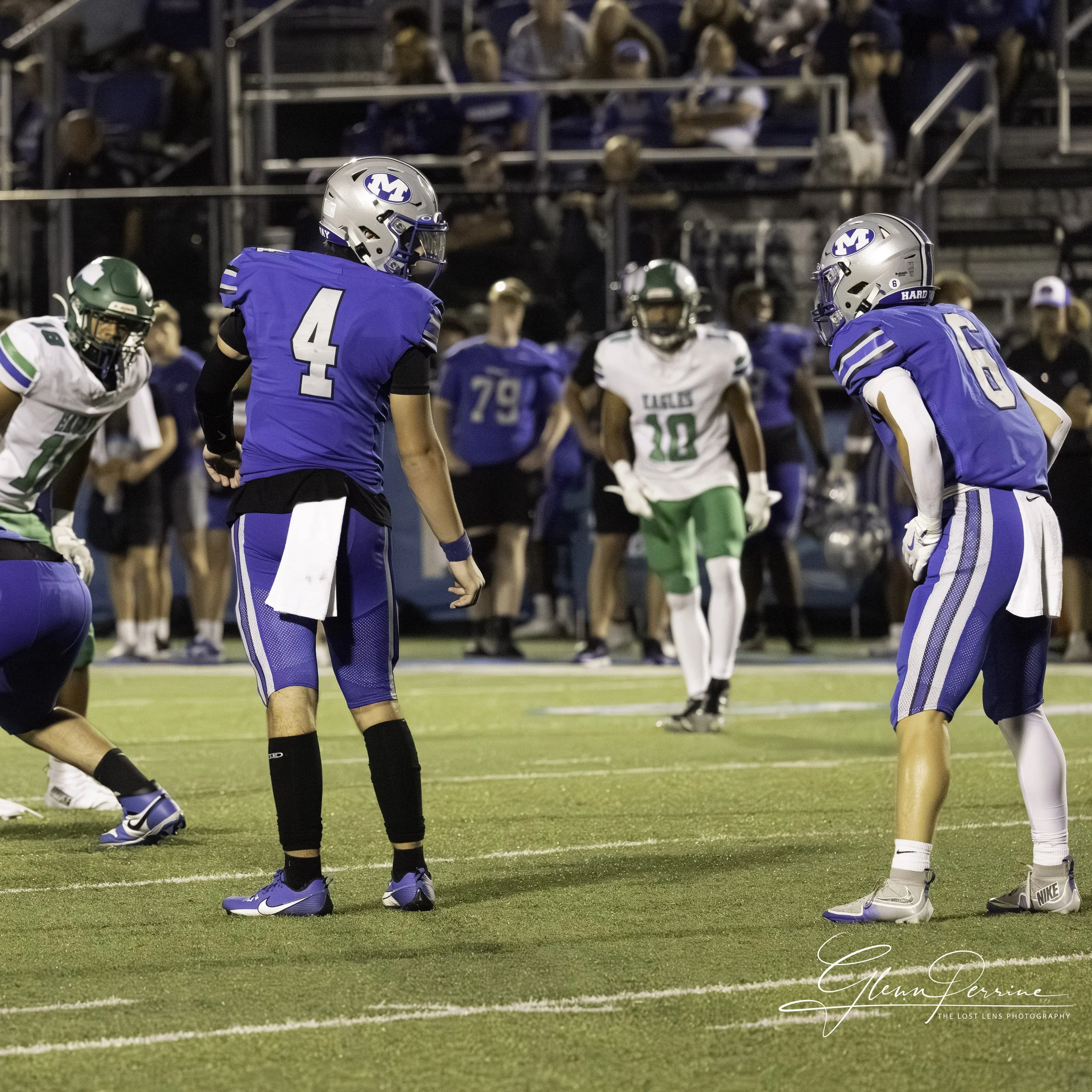 Football players in blue and green uniforms preparing for play on a football field at night, with spectators watching from the stands.