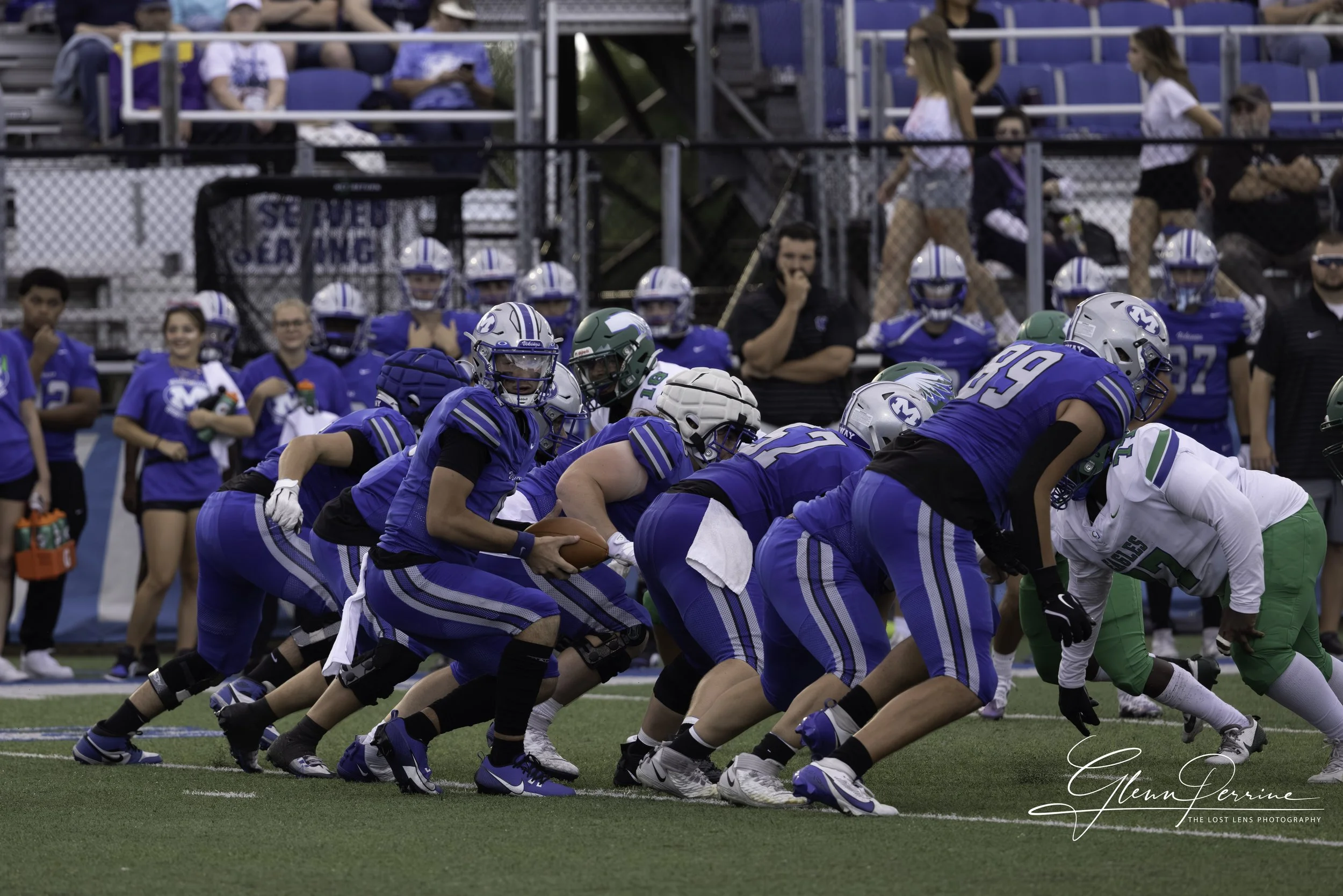 American football game with players in blue jerseys on offensive line ready to snap the ball, opposing team in white and green on defense, spectators in the stands and sidelines watching.