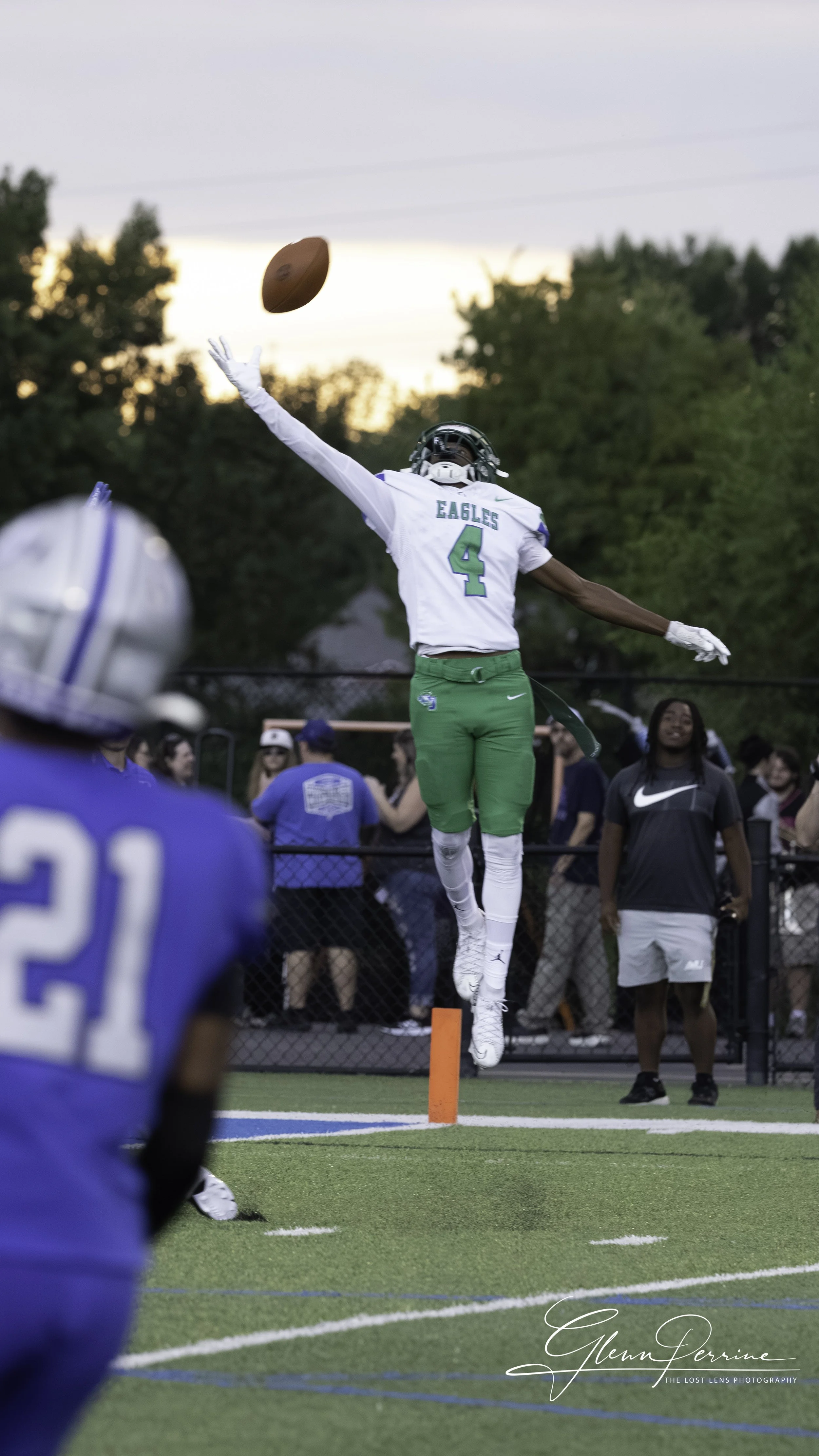 Football player in green and white uniform jumping to catch a football during a game, with other players and spectators in the background.