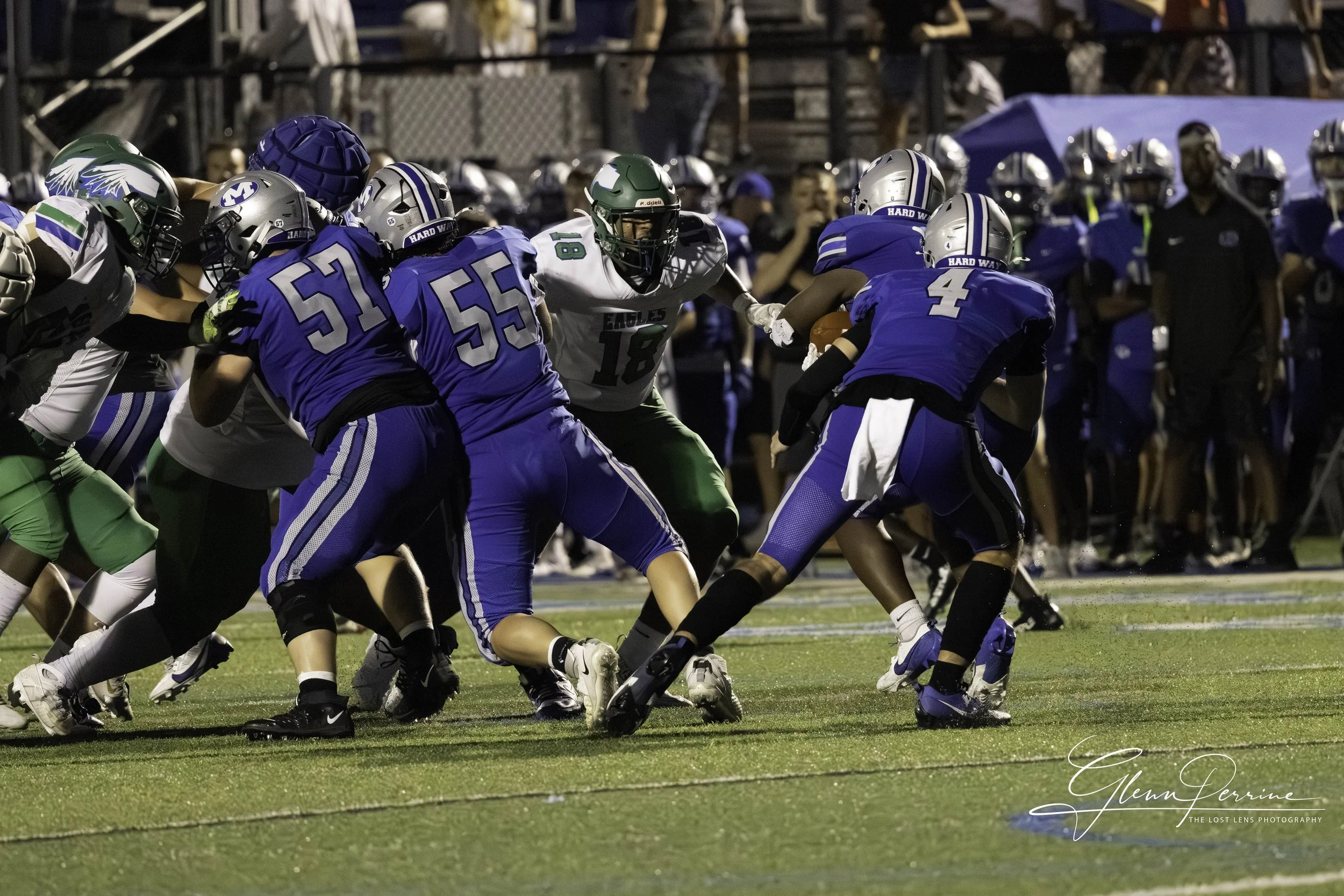 A youth football game with players from two teams, one in blue uniforms and the other in white and green, engaging in a play on a football field during evening, with onlookers in the background.
