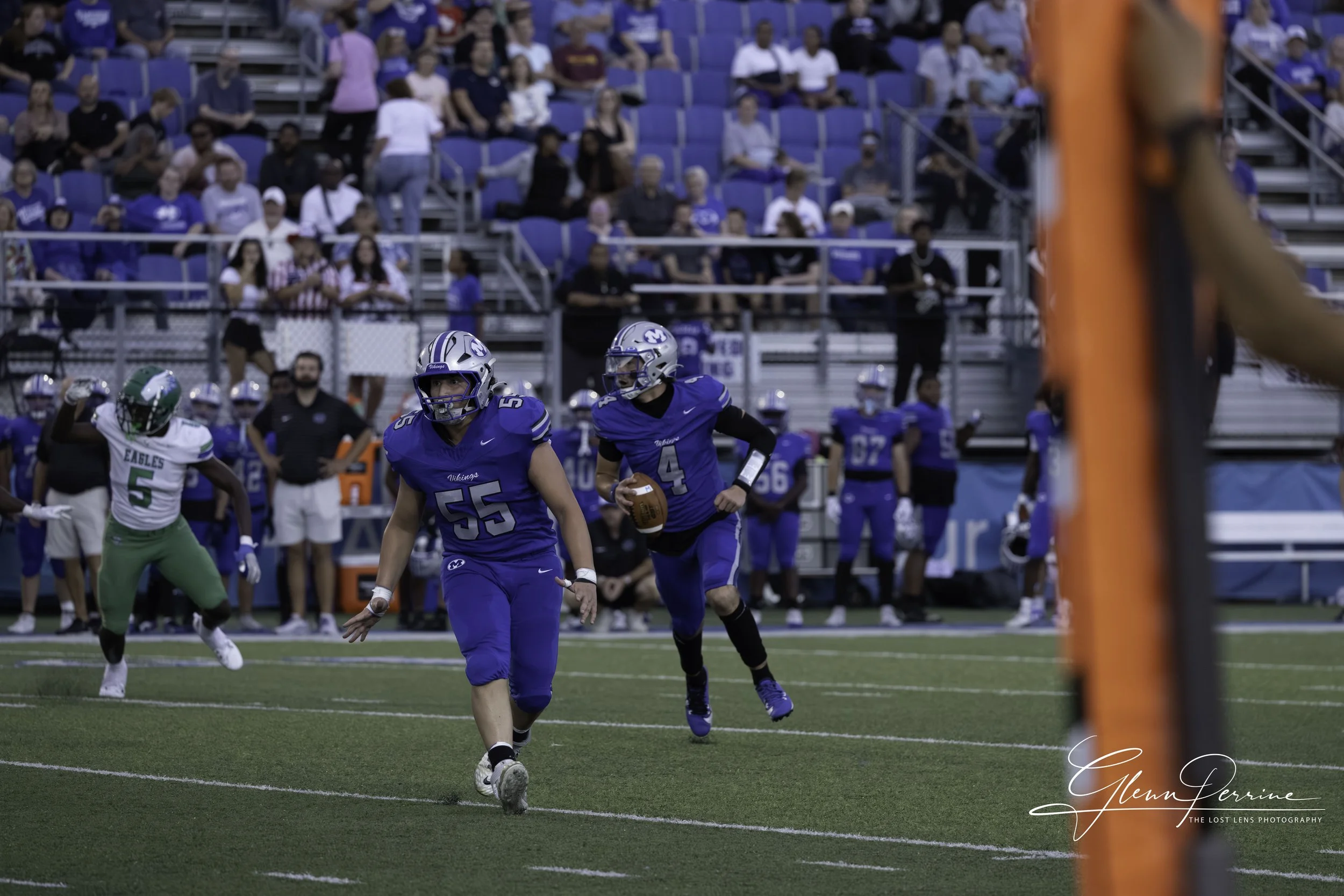 A football game with players dressed in blue and green uniforms. A player in blue is running with the football, while another blue player is behind him. An opposing player in a white and green uniform is nearby. The crowd is sitting on bleachers in t