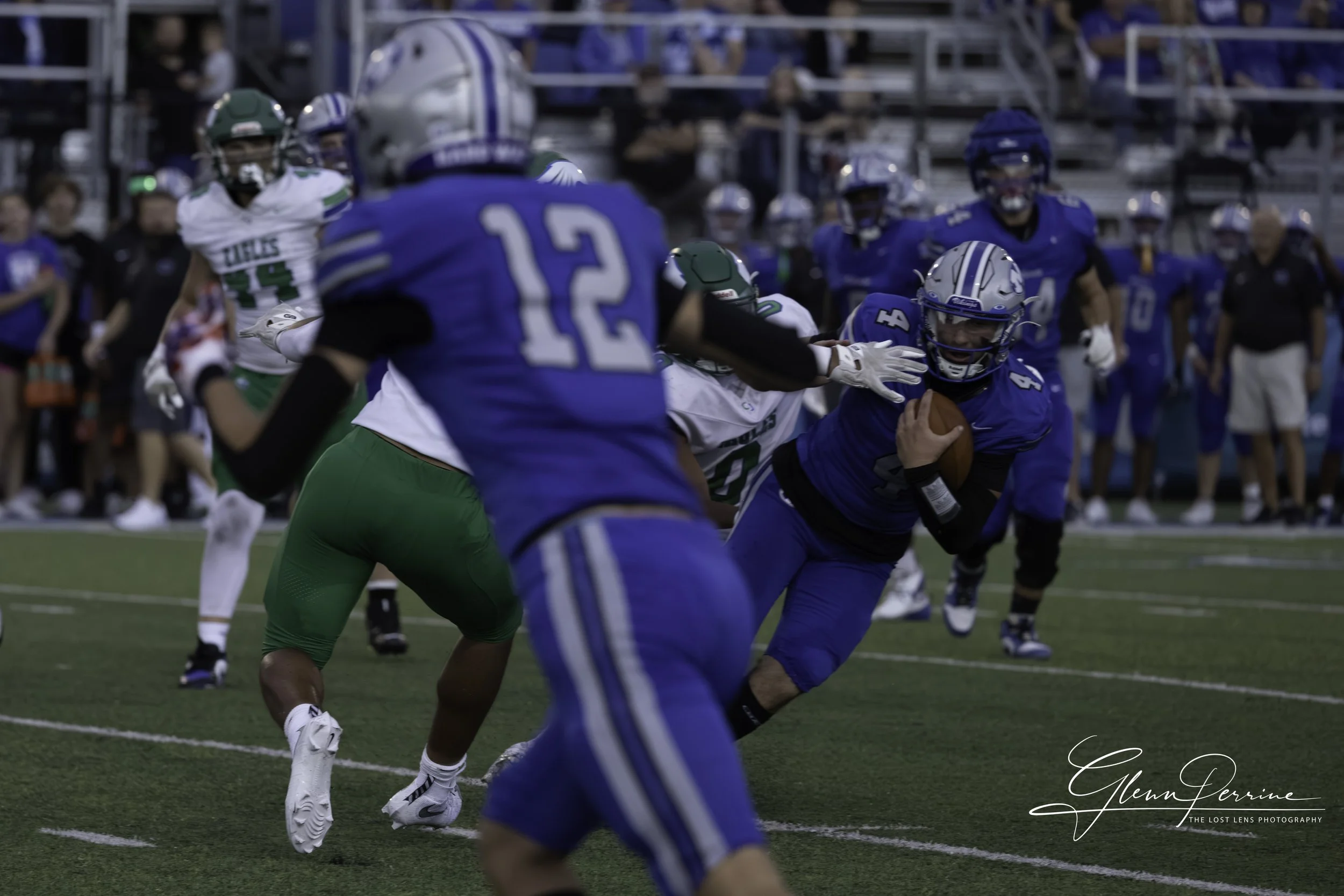A football game with a player in a blue uniform holding the ball and running, being tackled by a player in a white and green uniform. Several other players and spectators are visible in the background.