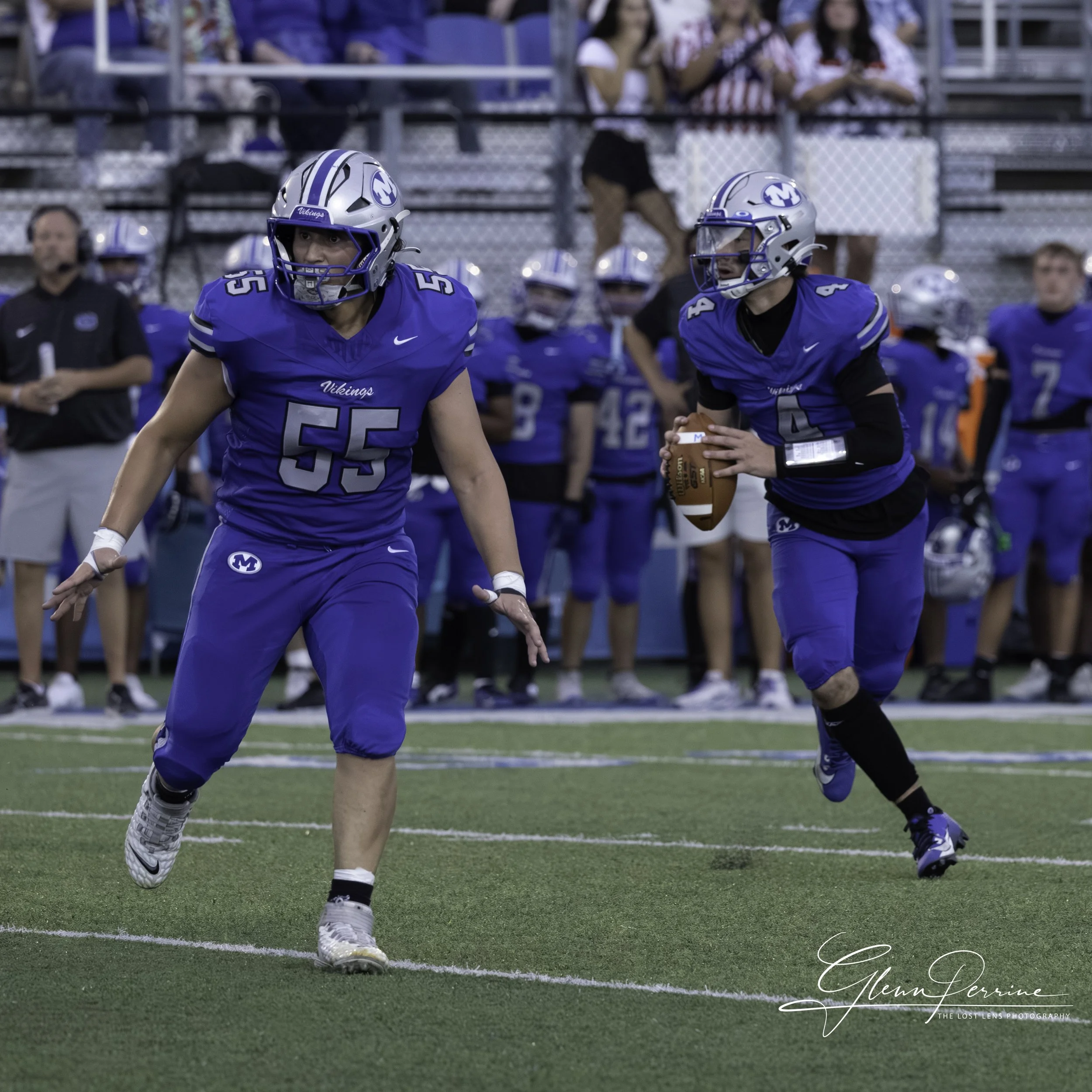 Two football players in blue jerseys and helmets, one holding a football ready to run, on a football field during a game, with fans and team members in the background.